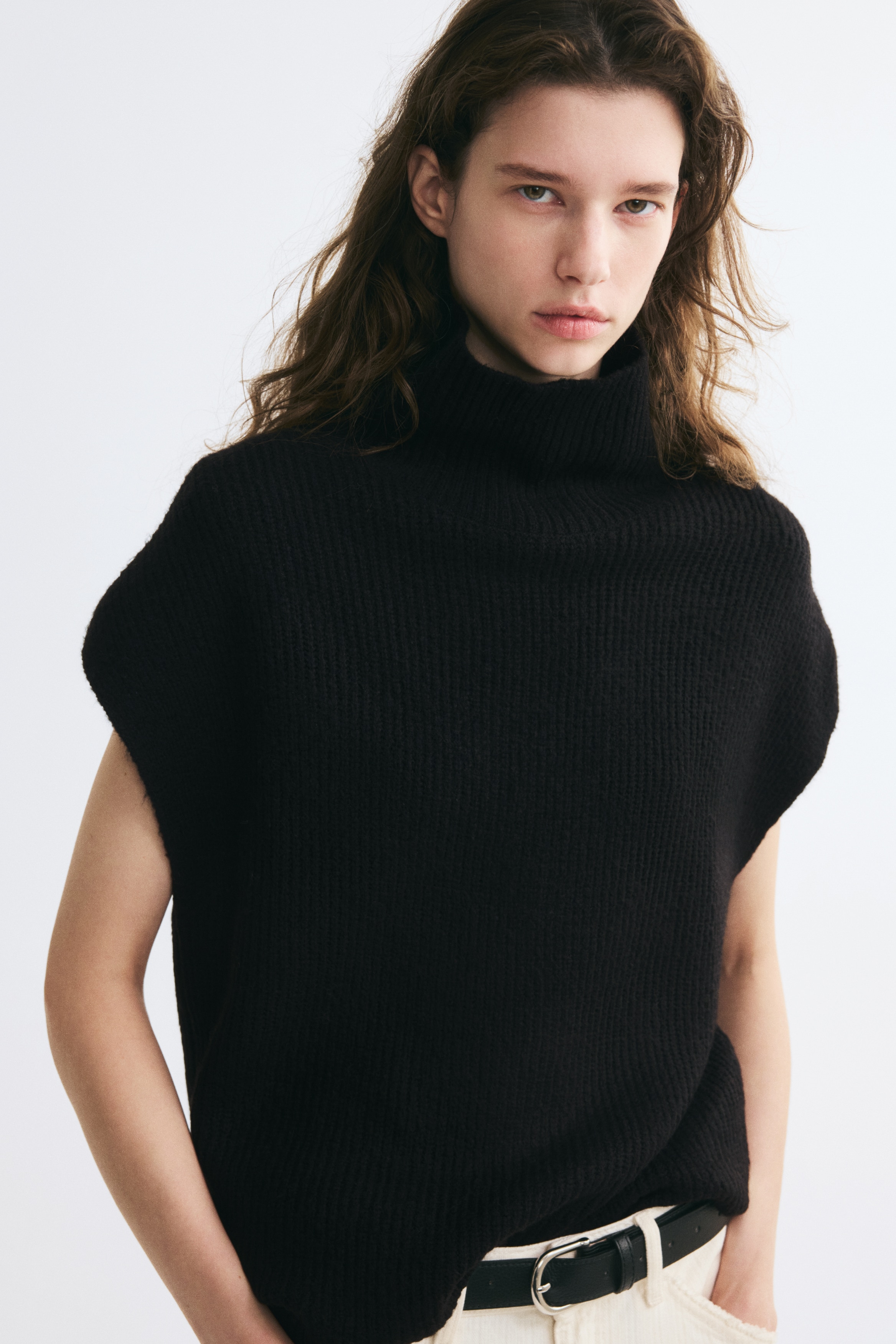 A black long jumper in a soft rib knit is facing front, showcasing a ribbed roll neck, short cap sleeves, and a straight hem. The garment features a consistent vertical ribbed texture throughout the body and collar.
