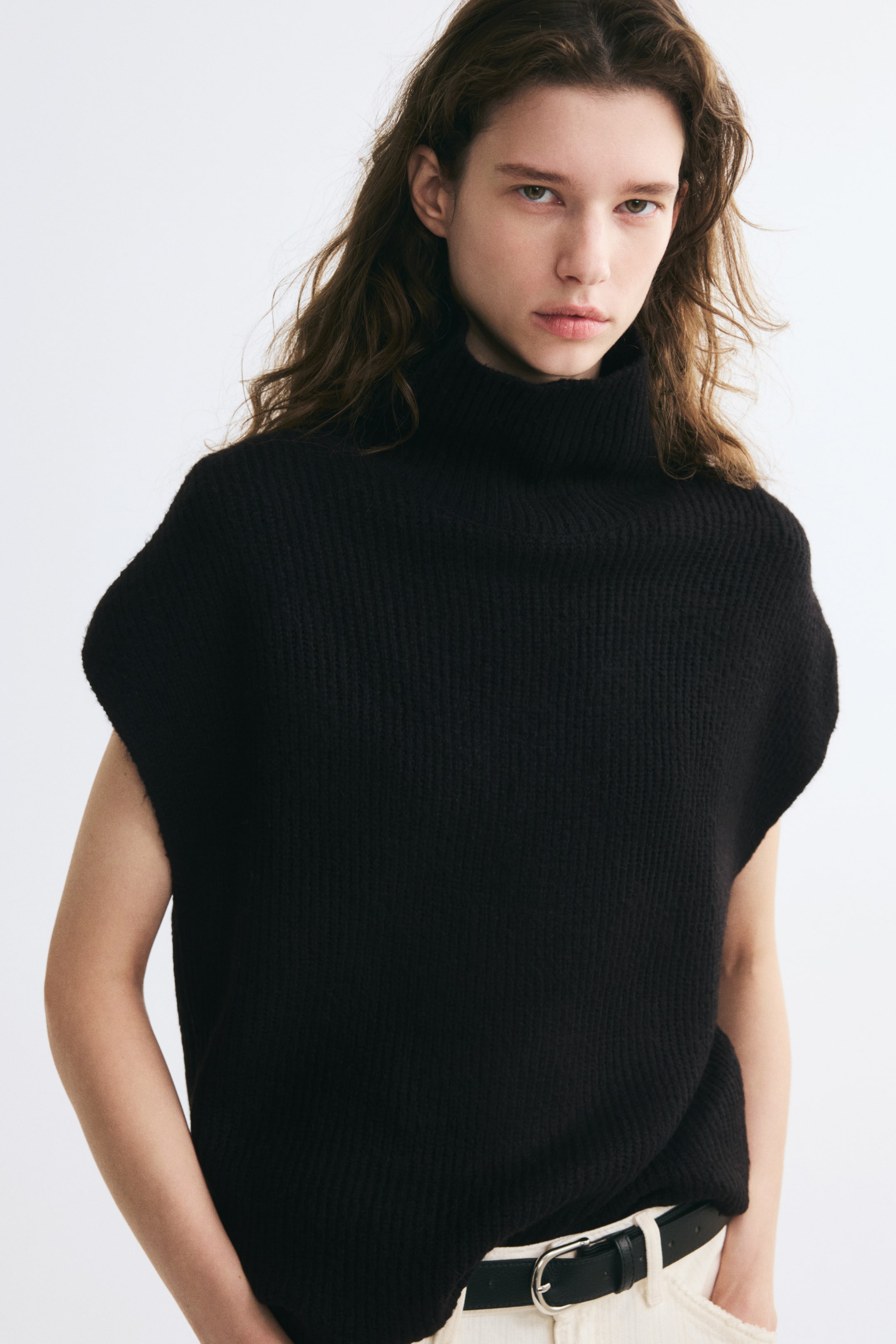 Größeres Bild ansehen: Ein schwarzer, gerippter Strickpullover mit Rollkragen und Cap-Ärmeln wird vom Model getragen. Der Pullover ist teilweise in hellfarbige Hosen gesteckt, die mit einem schwarzen Gürtel gehalten werden.