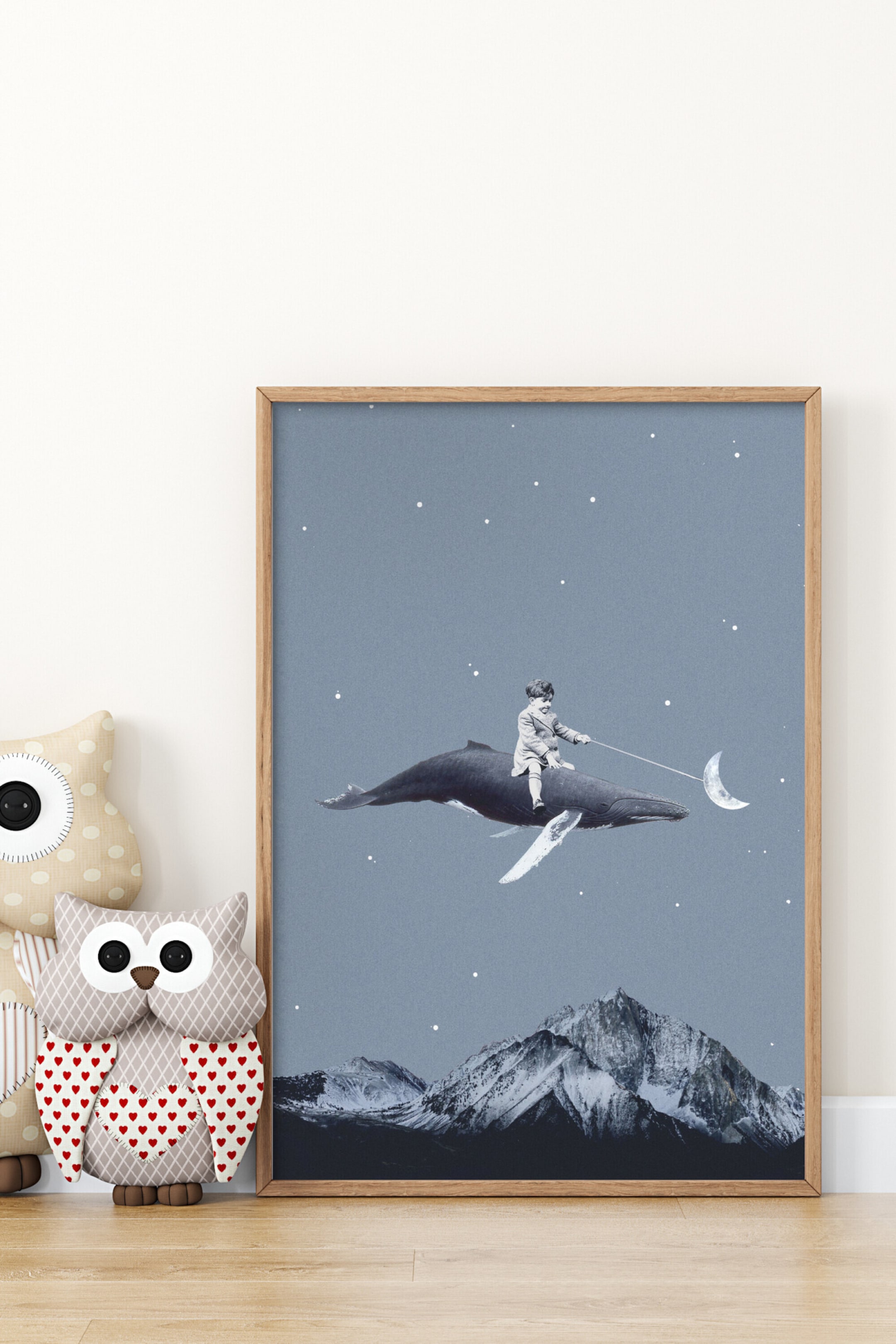 Visa större bild: En inramad konstprint visar en lekfull illustration av ett litet barn som rider på en val genom en stjärnbeströdd, dämpad blå himmel. Barnet håller i ett snöre som är fäst vid en halvmåne som en ballong, medan snötäckta bergstoppar ramar in kompositionens nederkant. Konstverket presenteras i en ljus naturfärgad träram.