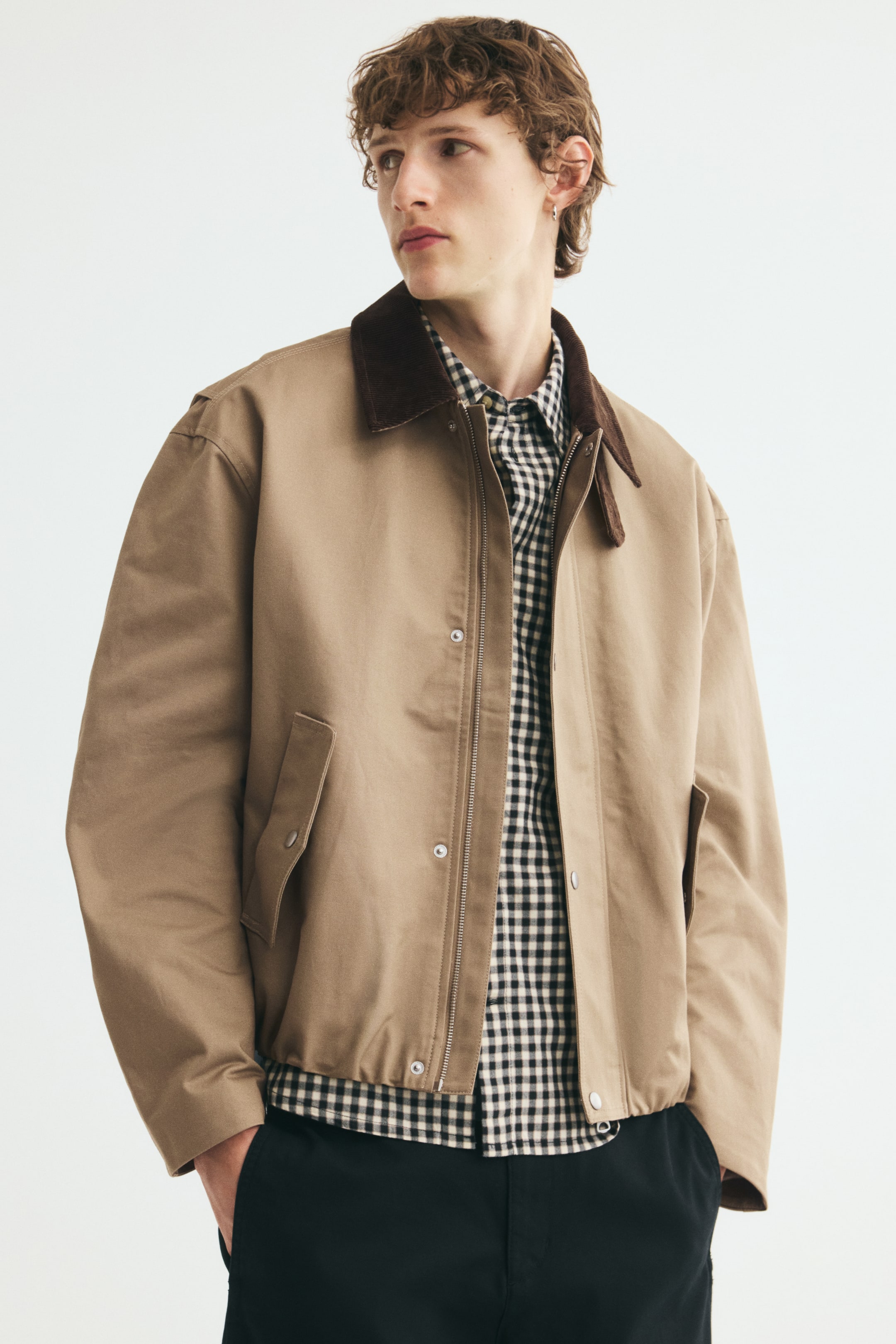 Xem ảnh lớn hơn: Một chiếc áo khoác chore bằng vải cotton twill màu be với cổ áo nhung corduroy nâu sẫm tương phản và dây cài cổ đang được người mẫu mặc. Chiếc áo khoác dáng mở phía trước được phối lớp bên ngoài áo sơ mi kẻ ô đen trắng và quần đen, làm nổi bật khóa kéo phía trước với nẹp chống gió cài nút bấm cùng các túi nắp bên hông.