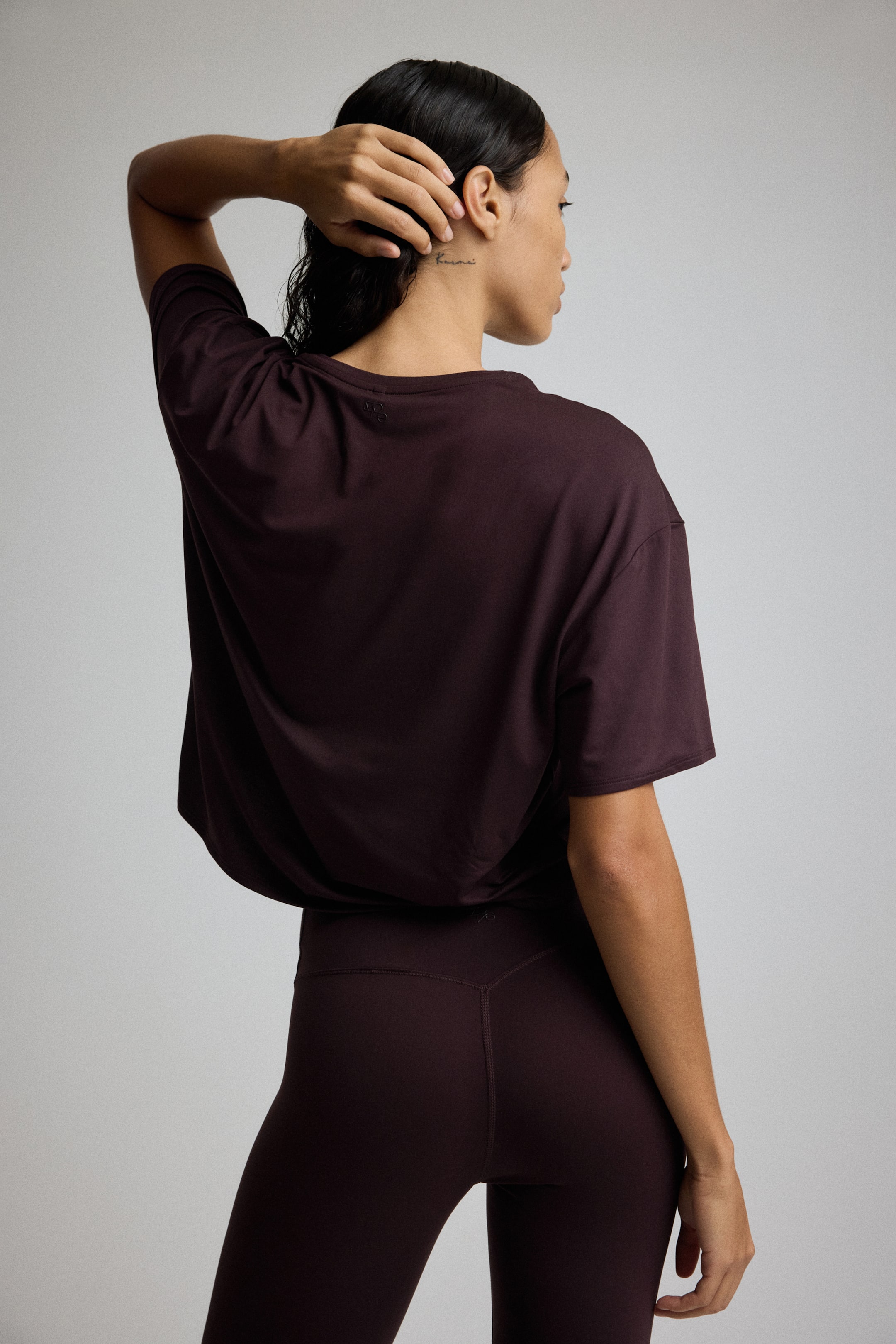 View larger image: A dark reddish-brown, short-sleeved t-shirt with a relaxed fit and a small logo on the upper back is being worn over matching high-waisted leggings that feature a V-shaped seam and a small logo on the back waistband, viewed from the back.