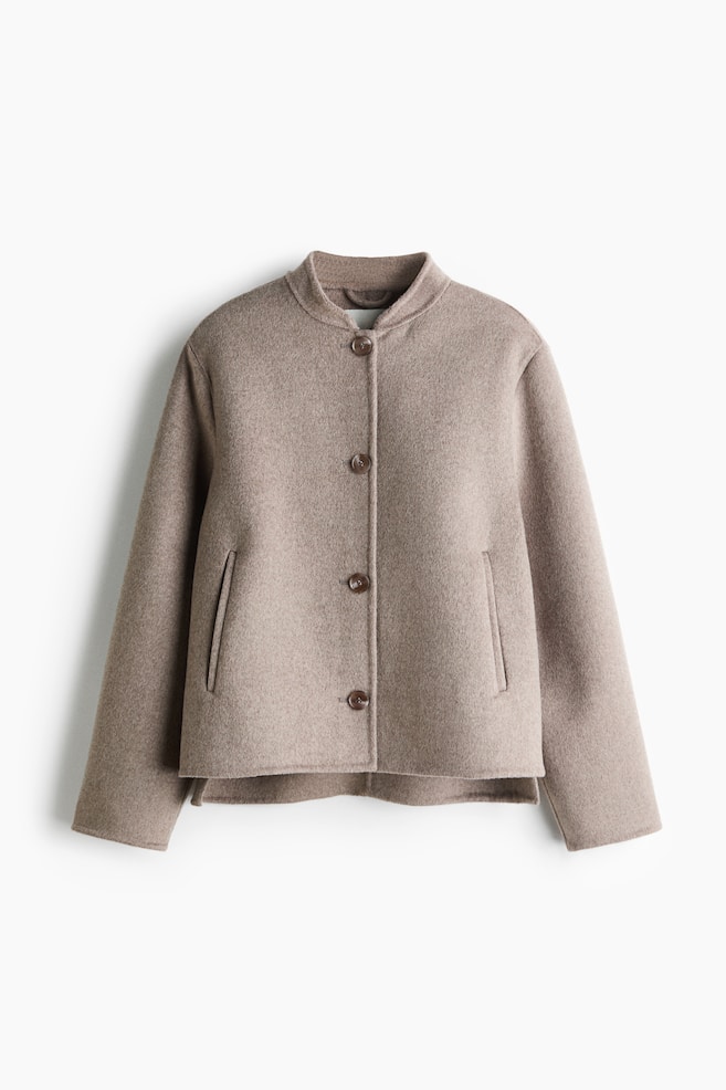 A taupe-coloured wool-blend jacket is shown from the front, showcasing a boxy, structured fit with a low band collar and a four-button front closure. The garment features long sleeves, vertical welt pockets, and a slightly stepped hem. - 1
