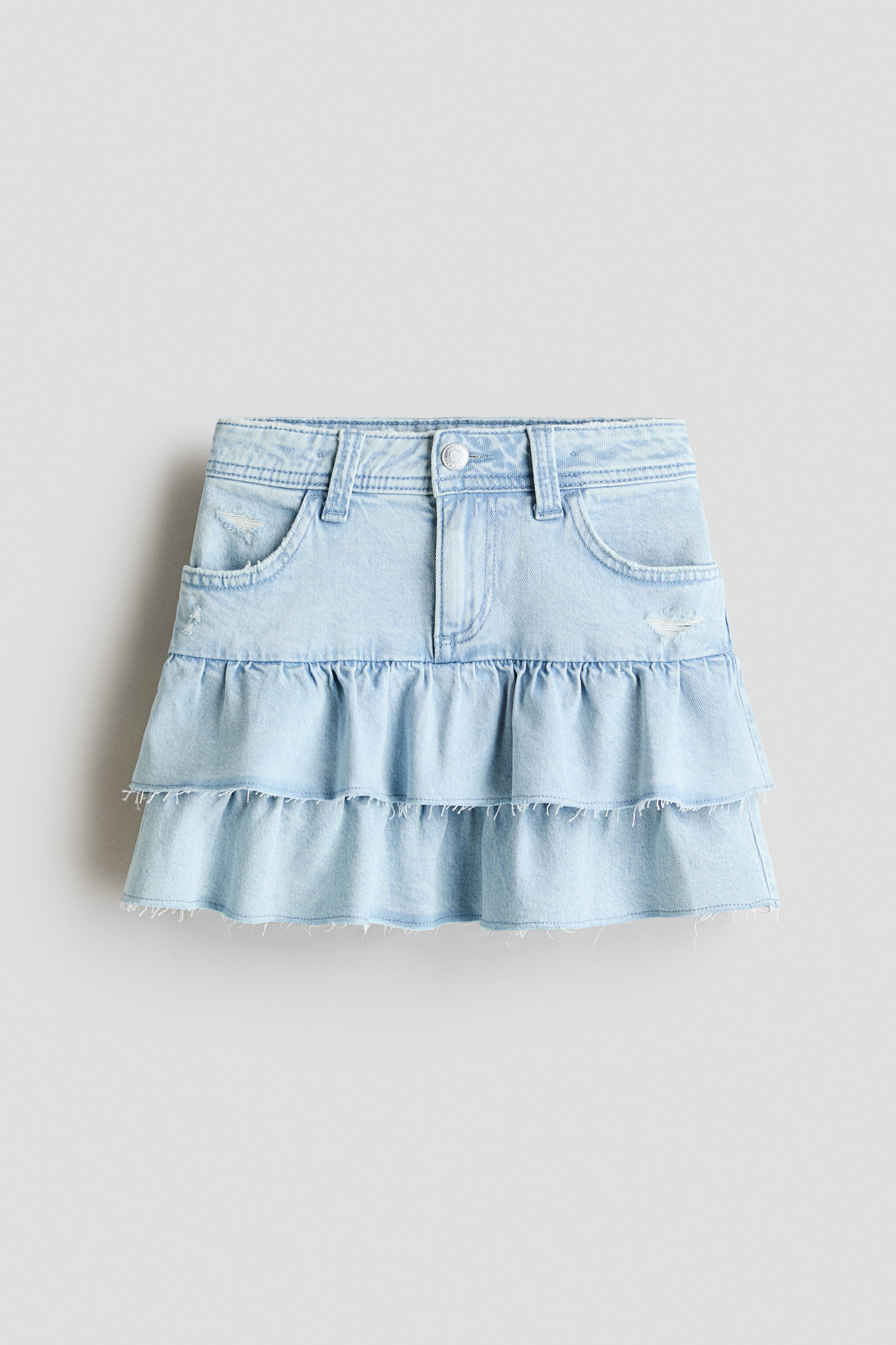 View larger image: "This light blue denim skirt showcases its front design, highlighting a waistband equipped with belt loops, a zip fly accompanied by a button closure, two curved front pockets adorned with distressed accents, and two ruffled tiers with raw edges forming the lower section."
