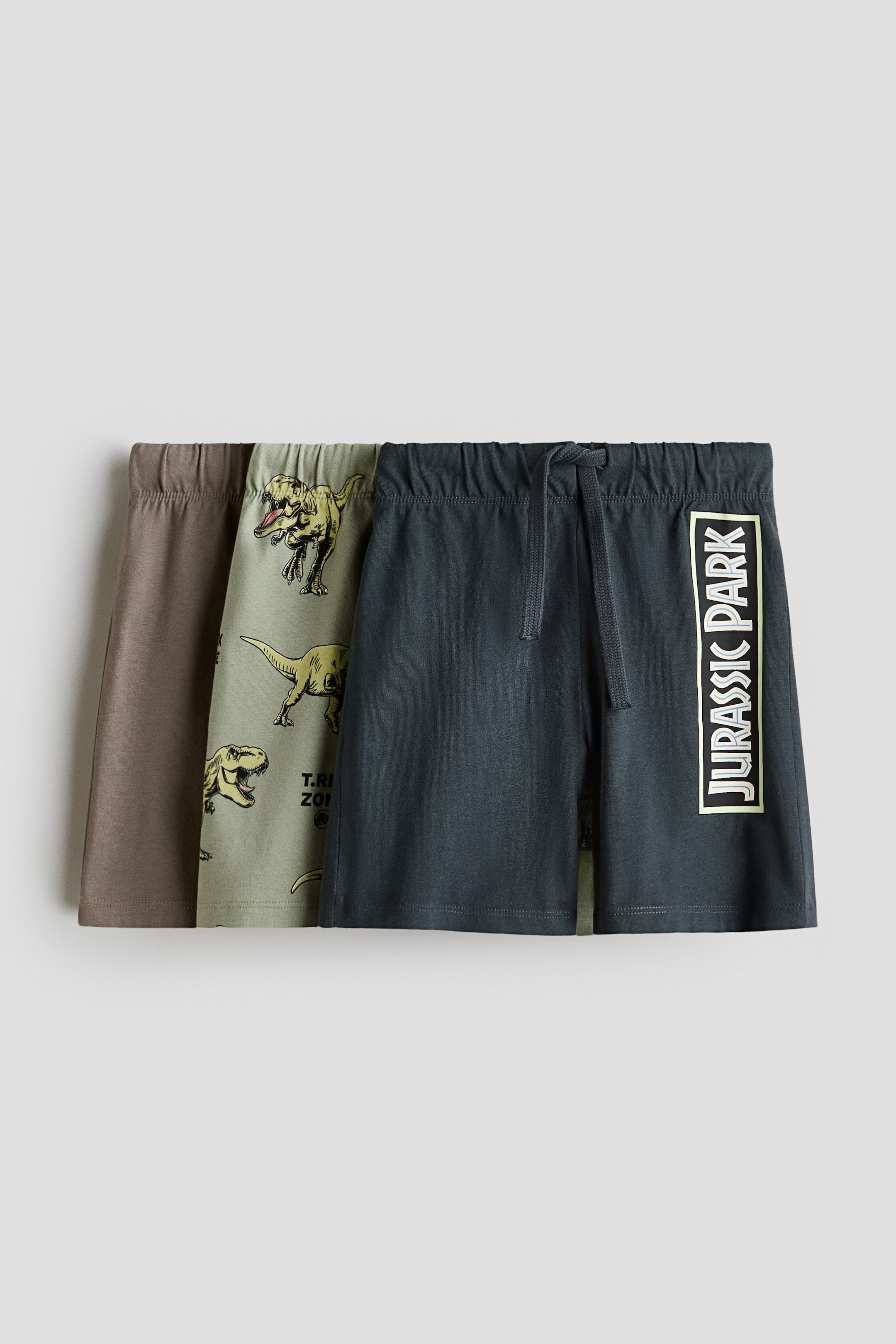 Ver imagen más grande: Tres pares de pantalones cortos presentan sus frentes, superpuestos. El par gris oscuro presenta un logotipo de Jurassic Park en la pierna y un cordón en su cintura elástica. El par verde claro muestra un estampado de dinosaurios, y el par marrón es liso.