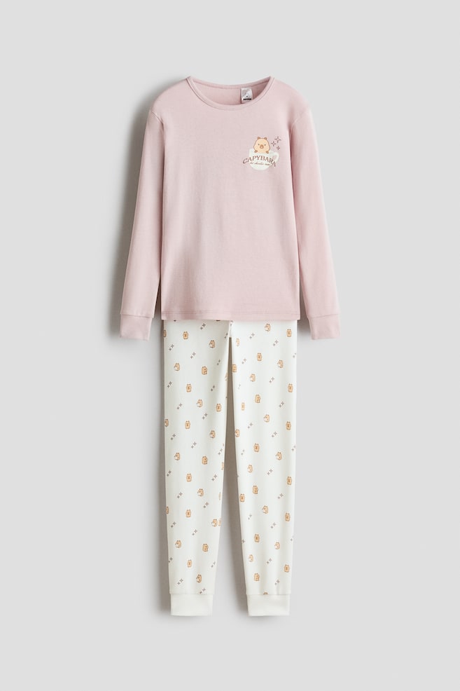 A front-facing pyjama set is featuring a light pink, long-sleeve top paired with off-white patterned pants. The top is displaying a round neckline with narrow trim, ribbed cuffs, and a small capybara graphic on the chest, while the pants are showing an all-over capybara print and ribbed hems. - 1
