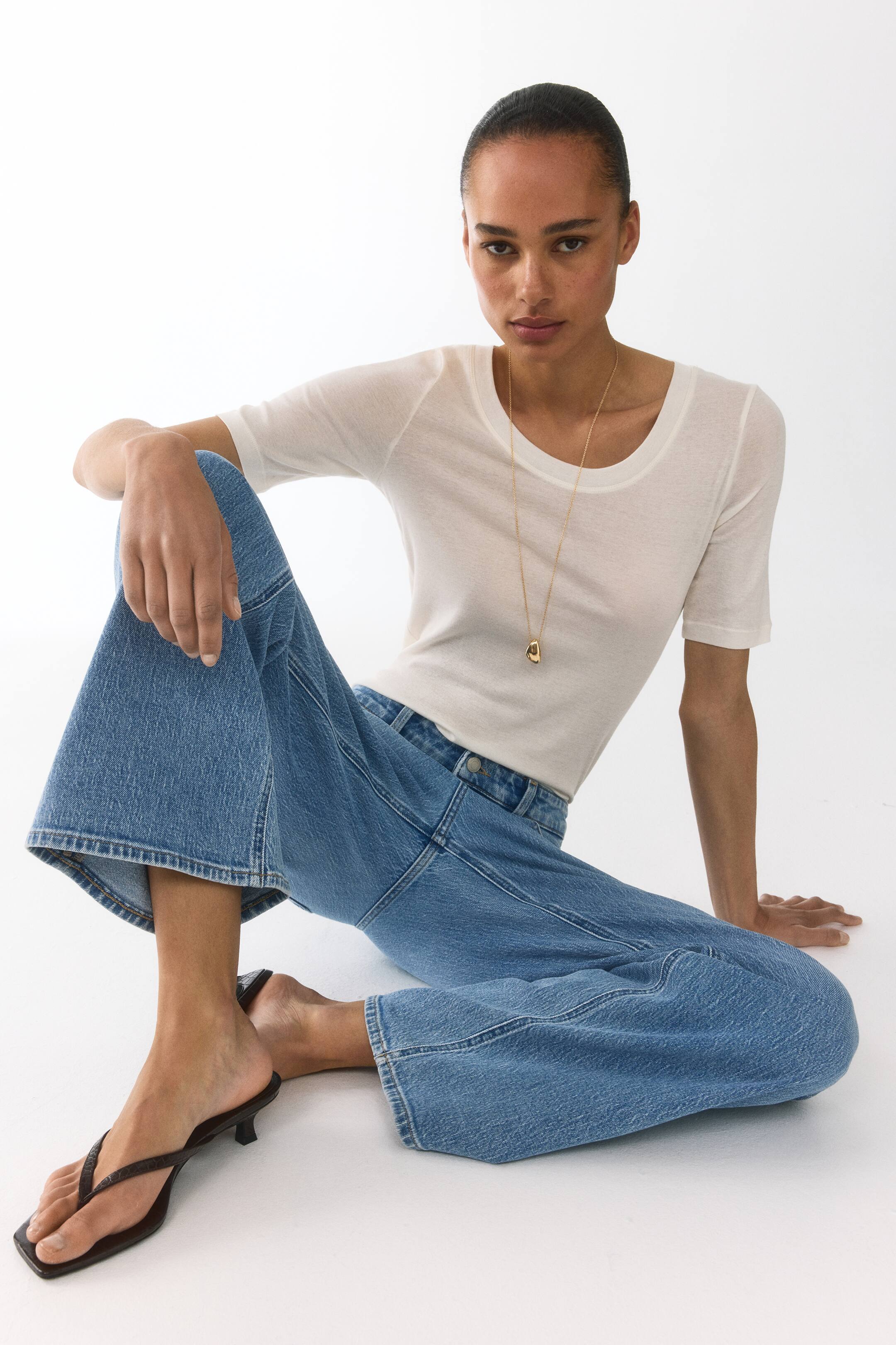 View larger image: A white, slightly sheer t-shirt features a scoop neckline and short sleeves, presented from a front-left angle. A gold pendant necklace drapes over its front.