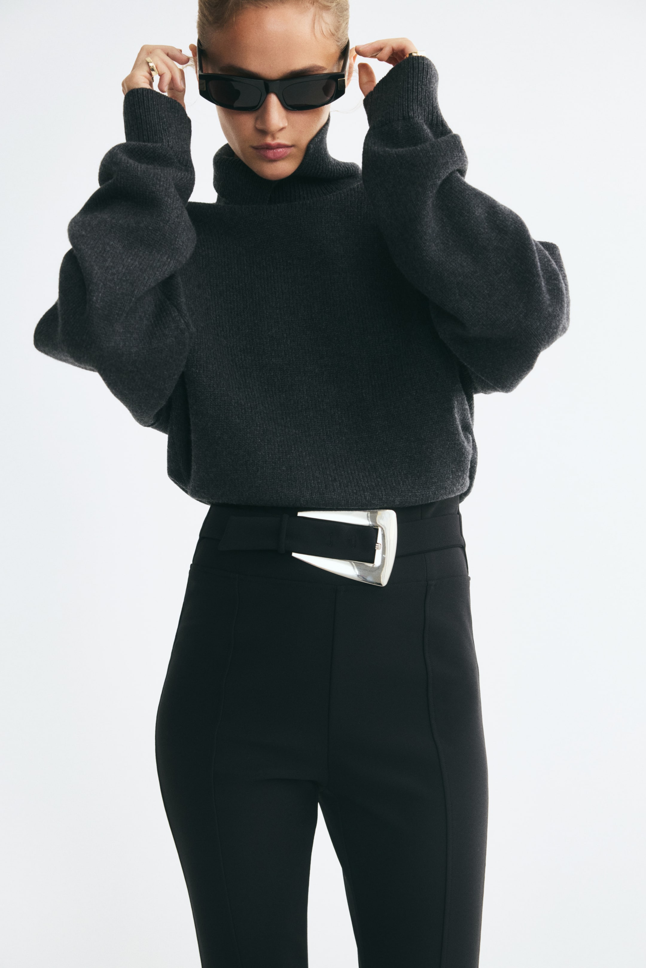 Ampliar la imagen: La modelo lleva leggings de jersey elástico negro, con pinzas cosidas al frente y un cinturón decorativo en la cintura con una gran hebilla metálica plateada asimétrica. Un suéter de cuello alto de punto en gris oscuro está metido dentro de la pretina alta, resaltando el detalle del cinturón y la hebilla.