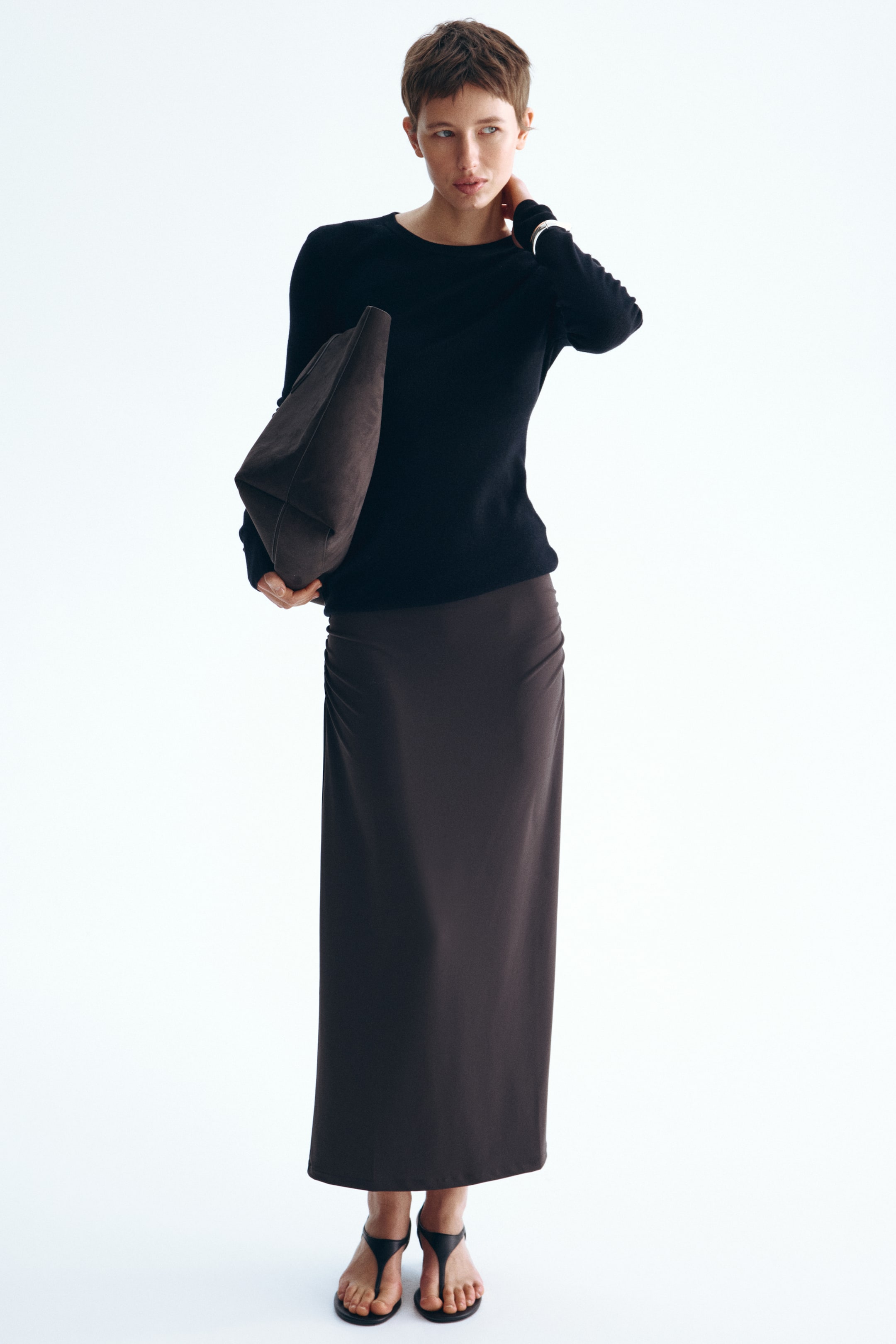 View larger image: A black long-sleeve crew-neck top fits snugly, featuring a silver bracelet on the left wrist, paired with a dark brown maxi skirt showcasing subtle ruching on its left side. A dark brown soft bag with a geometric folded design rests against the hip, while black toe-post flat sandals complete the look.