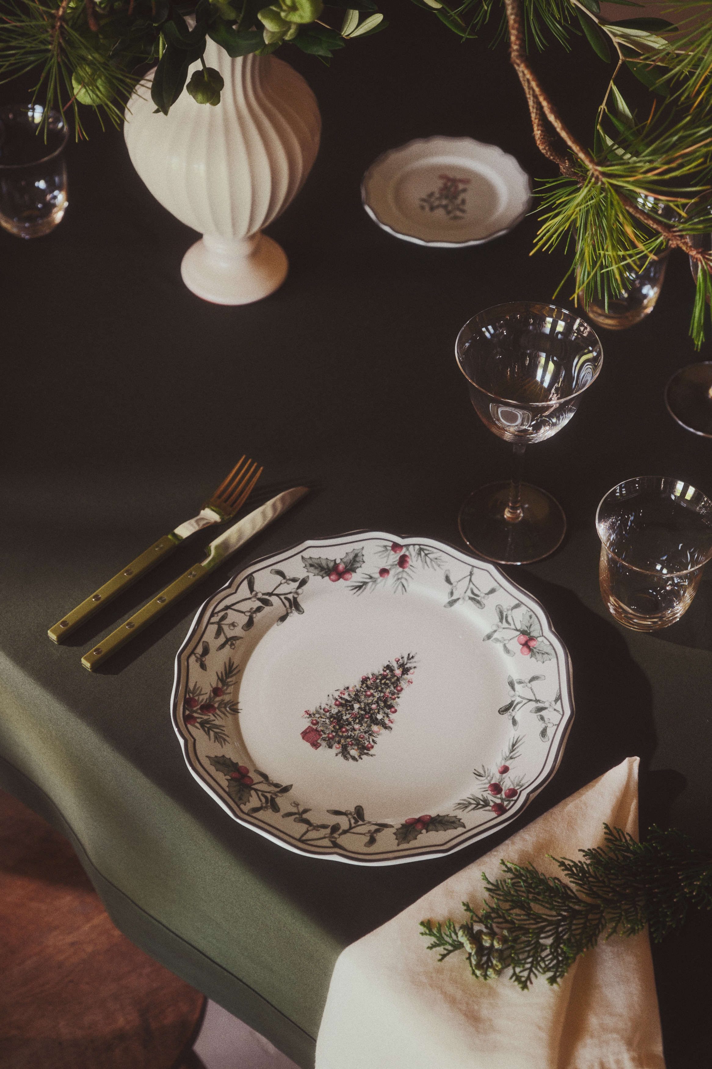 Four white ceramic plates are displayed in a festive design from a front-facing view, arranged in a two-by-two grid. Each plate features a scalloped edge with a thin dark rim, a central illustration of a decorated evergreen tree, and a border garland of green foliage and red berries.
