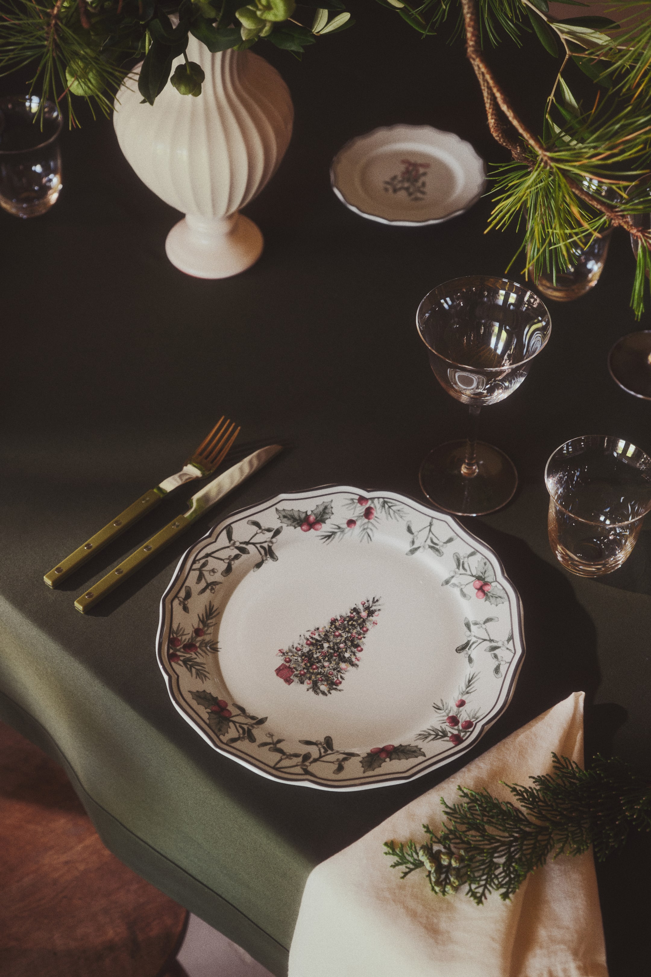 Voir une image plus grande: Une assiette plate en porcelaine présente un rebord ondulé aux accents sombres et des motifs imprimés festifs, avec une illustration centrale d'arbre décoré et une bordure de branches de houx aux baies rouges. L'assiette est montrée en plongée afin de mettre en valeur sa forme structurée et son motif détaillé.