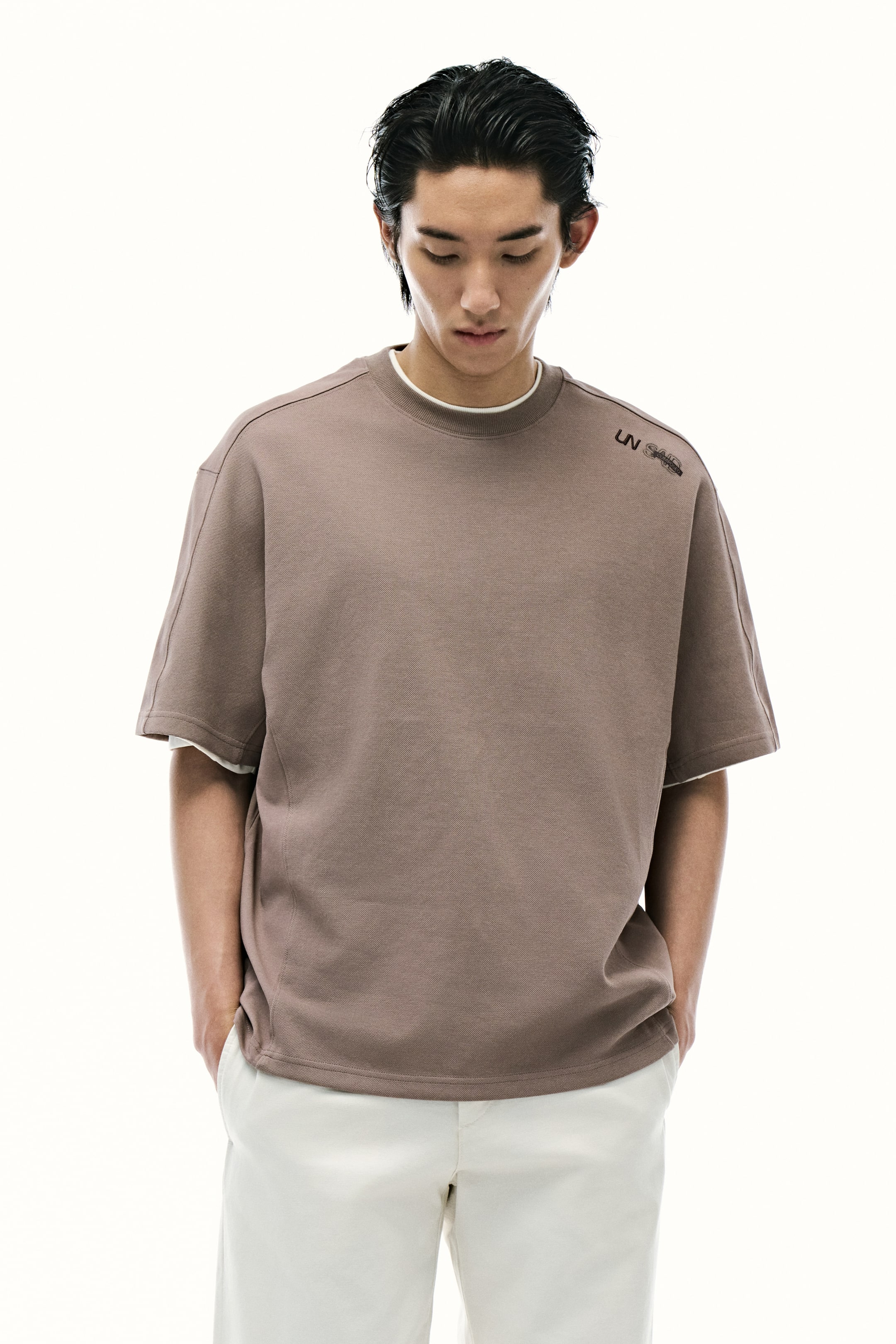 Xem ảnh lớn hơn: Một chiếc áo sweatshirt ngắn tay màu nâu rộng rãi đang hướng về phía trước, có cổ tròn và đường may vai nổi bật. Một hình ảnh đồ họa màu đen xuất hiện trên vai trái, và một lớp màu trắng được nhìn thấy phía dưới ở cổ áo và các khe tay áo.