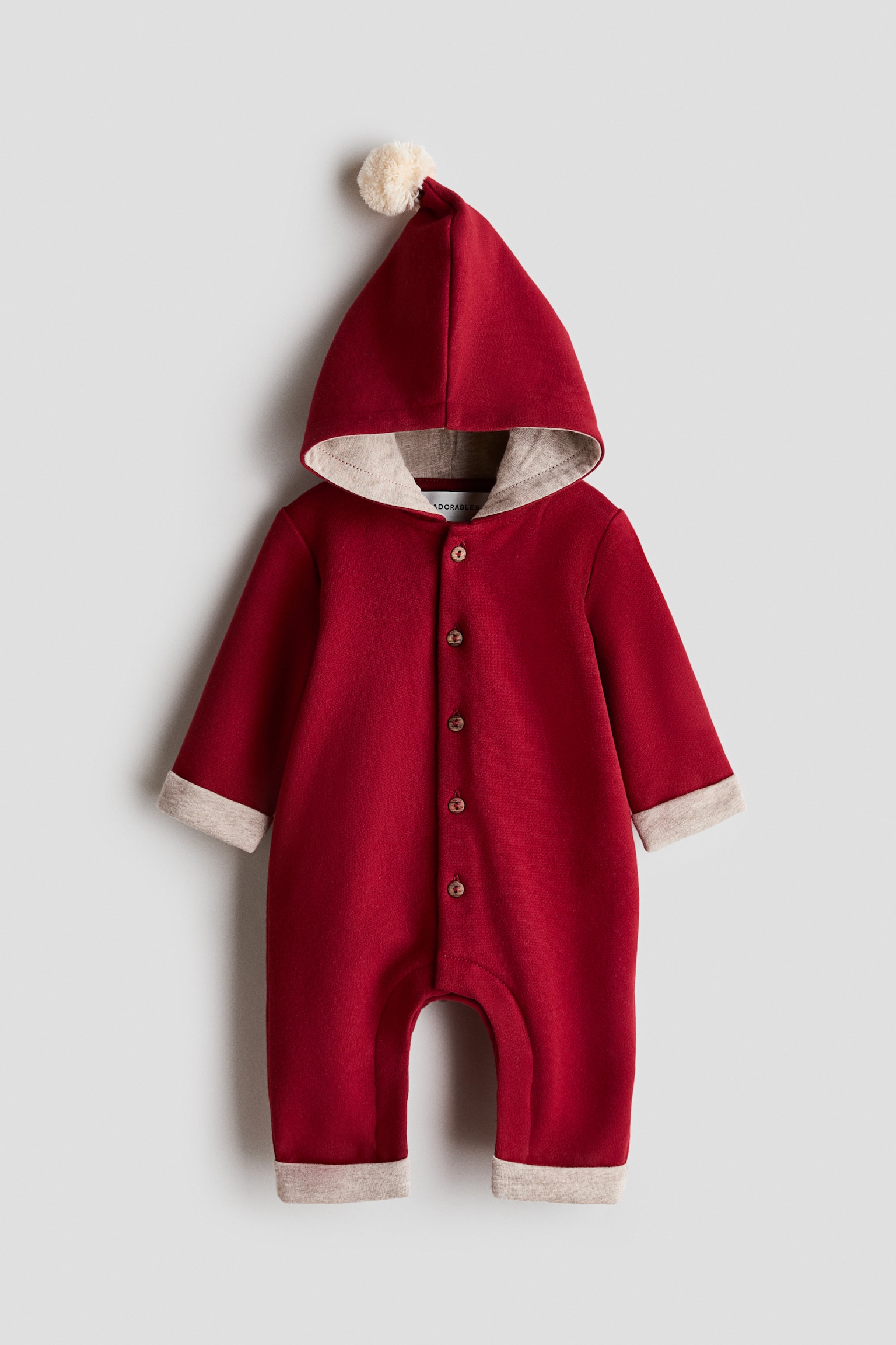 Una tutina per bebÃ© rossa con cappuccio Ã¨ distesa in piano, rivolta frontalmente, e mette in risalto un cappuccio a punta sormontato da un pompon color crema. La tutina presenta una chiusura centrale con bottoni, con cinque bottoni in legno, e una fodera beige a contrasto visibile allâinterno del cappuccio, oltre che sui risvolti delle maniche e dei polsini delle gambe.