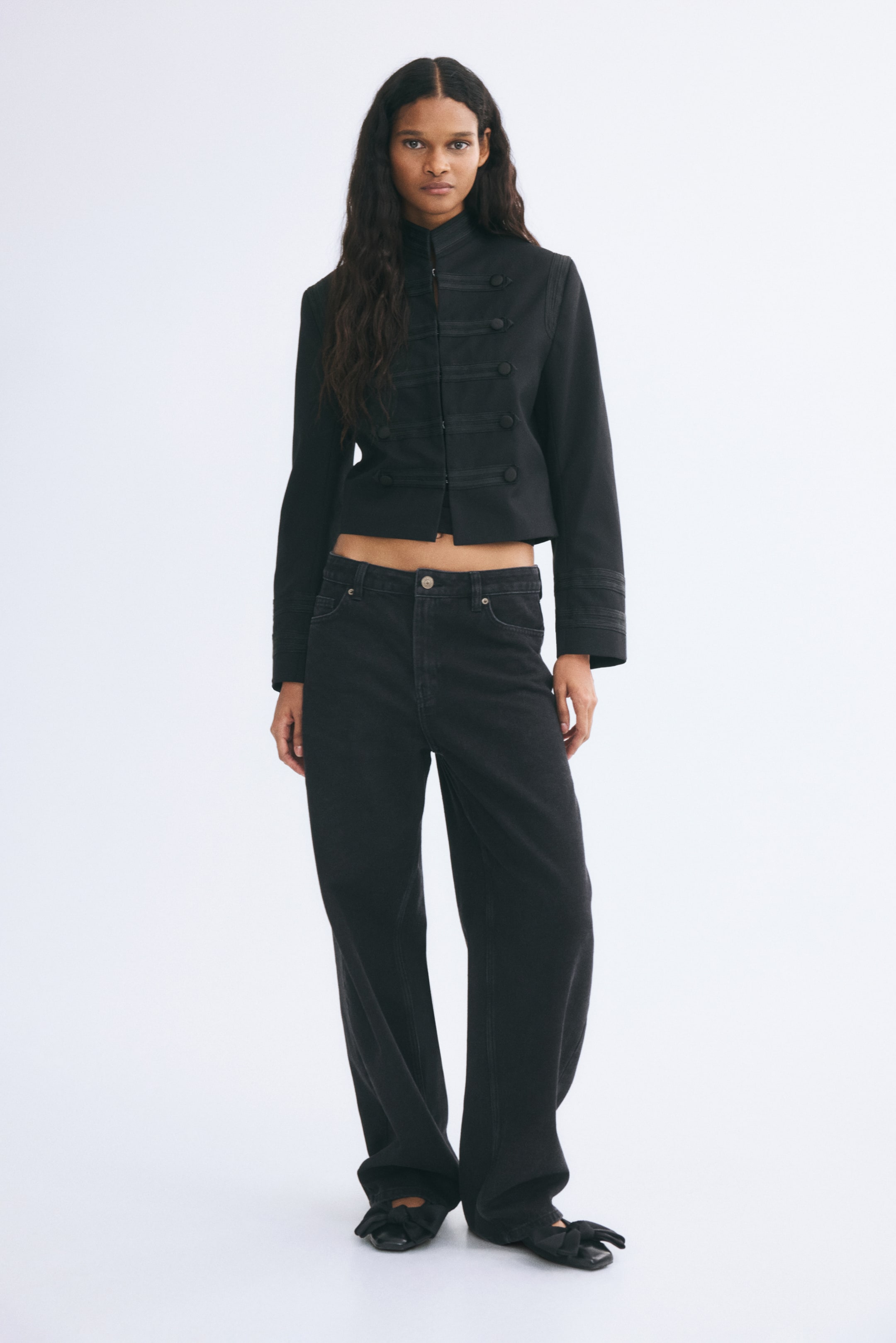 Xem ảnh lớn hơn: Quần jeans denim đen 5 túi với cạp vừa và ống quần cong, phồng đang được người mẫu mặc. Quần được phối cùng áo khoác đen tay dài dáng lửng với chi tiết nút kiểu quân đội và giày bệt màu đen.