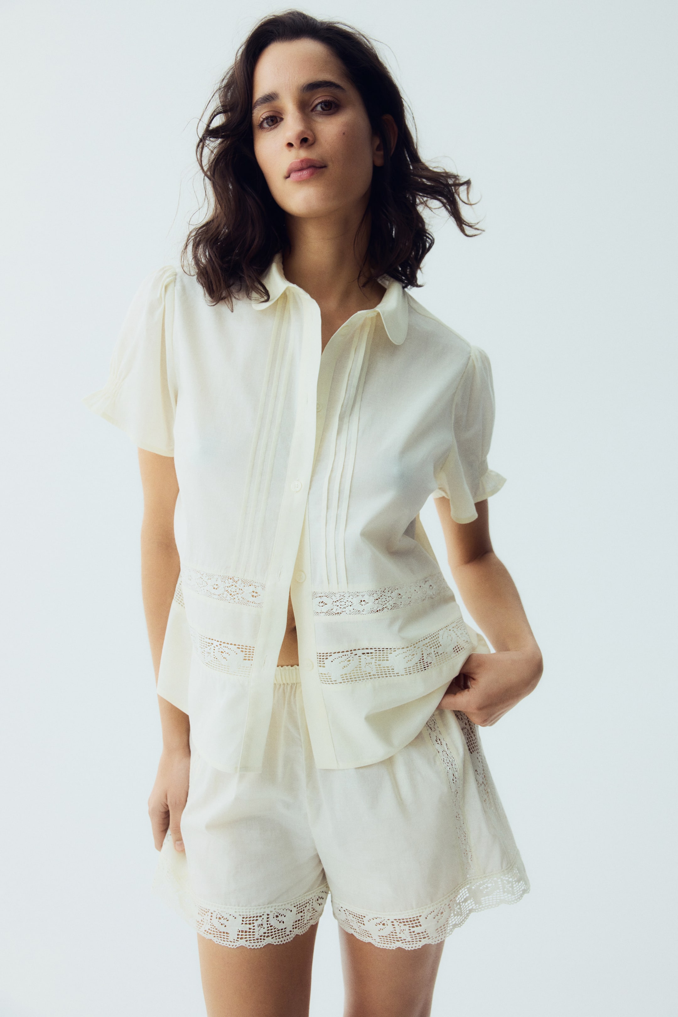 View larger image: A slightly angled front view showcases an off-white two-piece set. The short-sleeved collared top features vertical pin-tucks flanking the partially unbuttoned front, complemented by horizontal lace inserts across the mid-section and puffed sleeves. The matching shorts highlight an elastic waistband and lace trim along the leg openings.