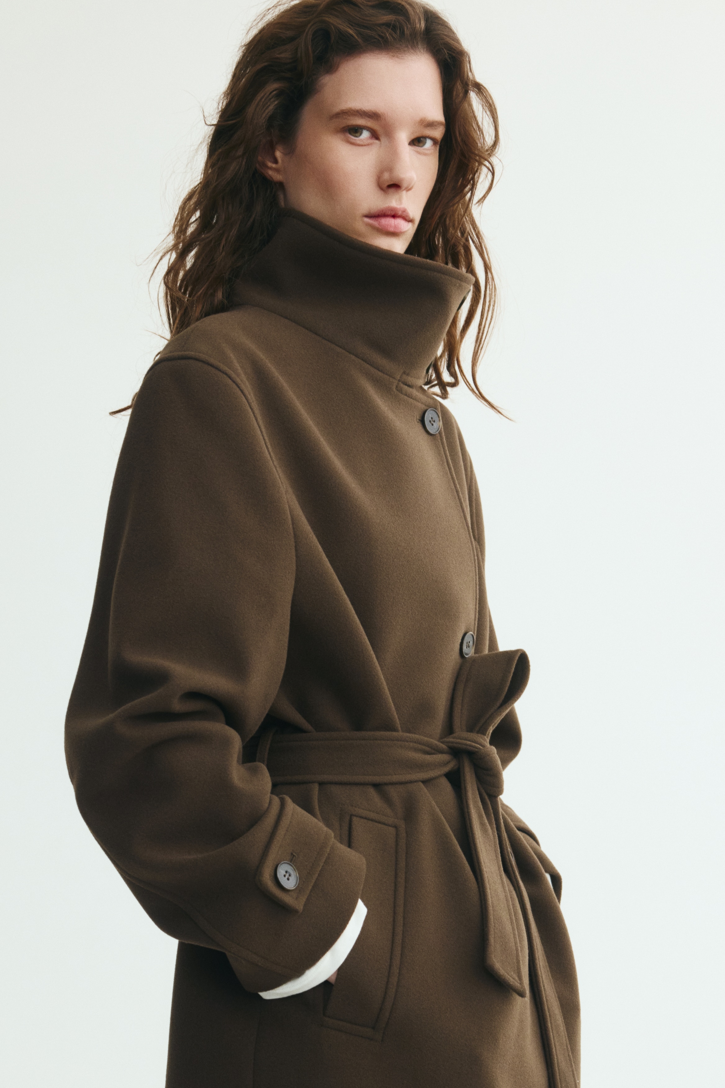 Een lange, oversized donkerbruine jas in een gestructureerde geweven stof wordt van voren getoond, met een hoge, geknoopte opstaande kraag en een asymmetrische knoopsluiting. De jas is gestyled met een bijpassende stoffen ceintuur die in de taille is gestrikt, schuine paspelzakken en manchetten met knoopsluiting.