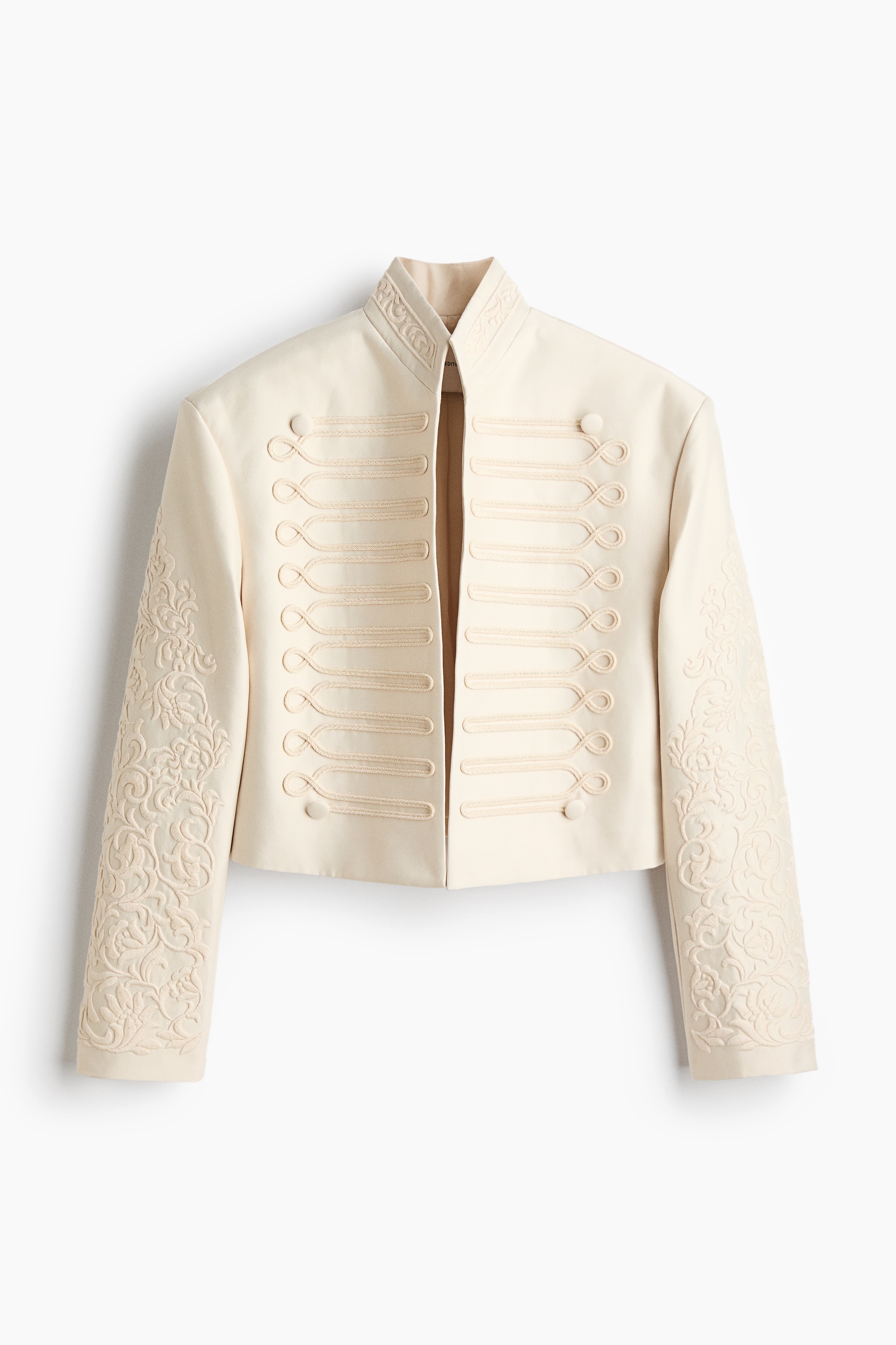 Una chaqueta corta de sarga color crema con cuello alzado, detalles bordados en las mangas y adornos decorativos de cordÃ³n en la parte delantera, llevada abierta por la modelo. La chaqueta se combina con un top de encaje semitransparente y se coordina con pantalones blancos.