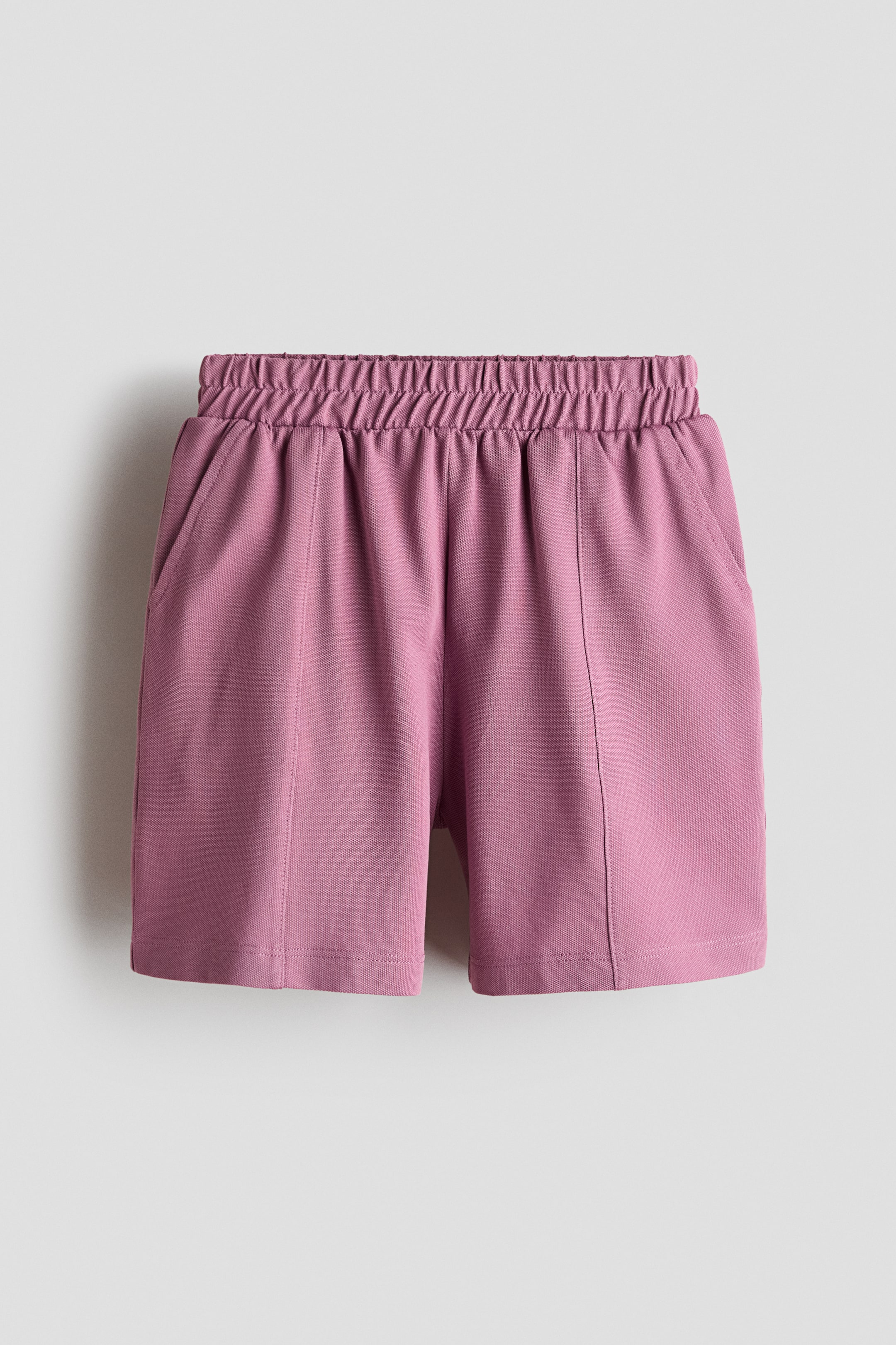 Xem ảnh lớn hơn: Một chiếc quần shorts màu hồng phấn đang được chụp chính diện, với cạp chun nhún và túi xéo hai bên. Quần có các đường may dọc nổi chạy xuống mặt trước mỗi ống, đến phần gấu được hoàn thiện gọn gàng.