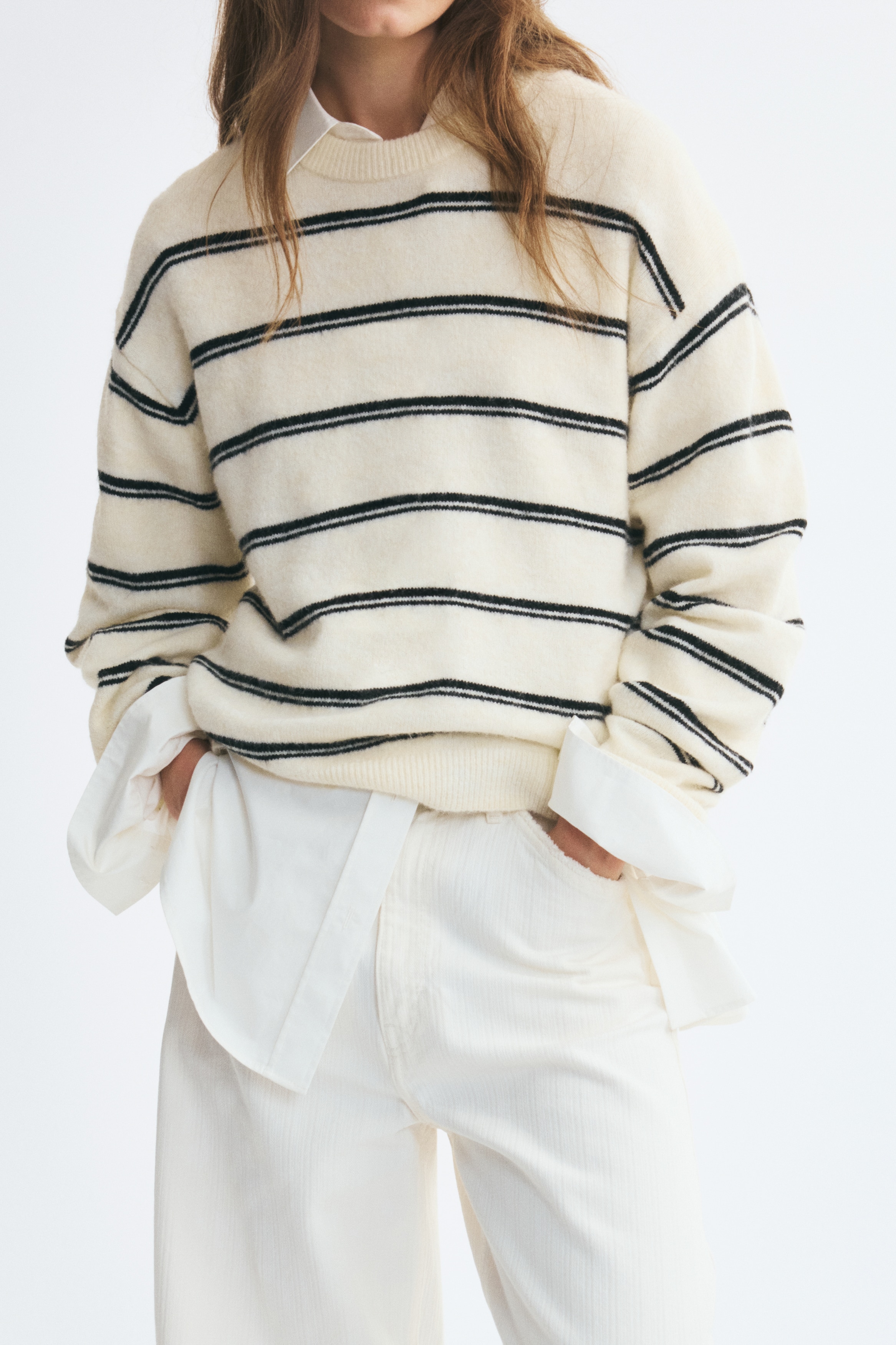 A cream-colored knit sweater is shown from the front, featuring horizontal black double stripes across the body and long sleeves. The sweater is styled with a round, rib-trimmed neckline, dropped shoulders, and ribbing at the cuffs and hem.