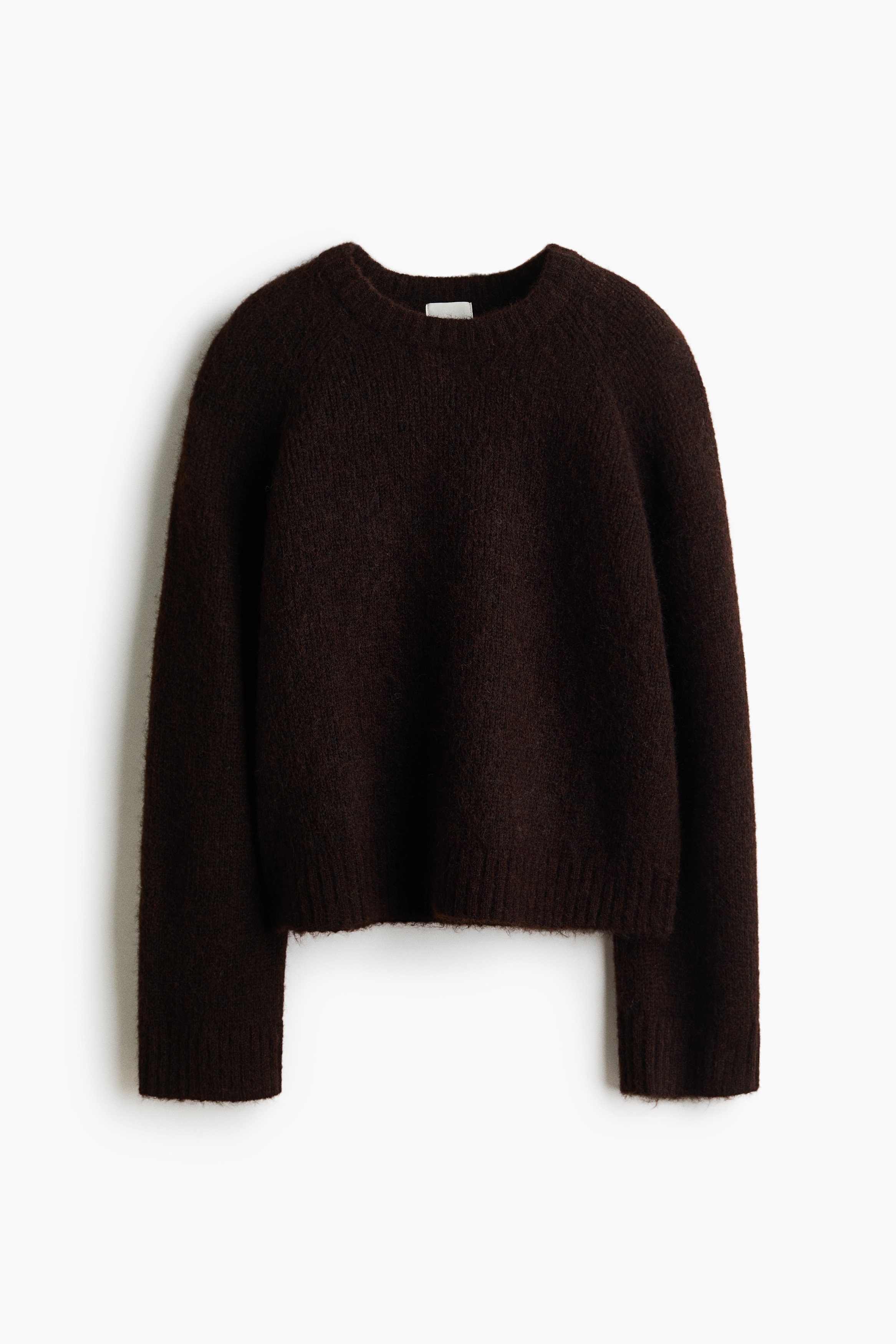 Een oversized, donkerbruine trui van zachte knit wordt door het model gedragen, met een ronde, geribde halslijn, lange mouwen met geribde boorden en een geribde zoom. Het model houdt met Ã©Ã©n hand de zijkant van de trui ter hoogte van de taille vast en trekt deze iets samen, waardoor de tailleband van een donkerleren broek zichtbaar wordt.