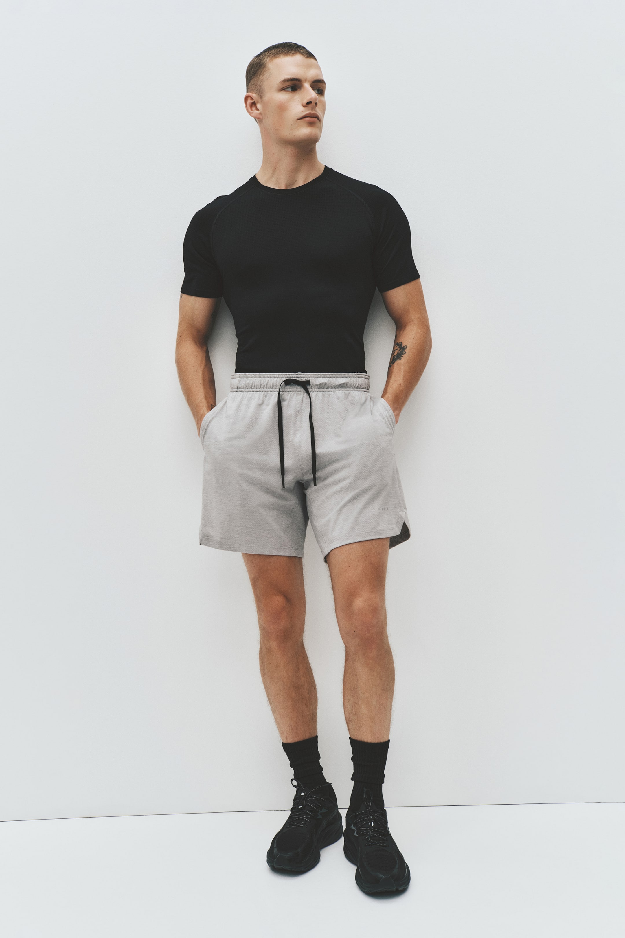 View larger image: The product showcases a black short-sleeved athletic t-shirt with visible raglan seams and a crew neck, designed for a snug fit. Light grey athletic shorts feature an elastic waistband, a black drawstring, side pockets, and a dark grey contrasting lower side panel, providing a relaxed fit. Black crew socks and black lace-up athletic sneakers complete the look.