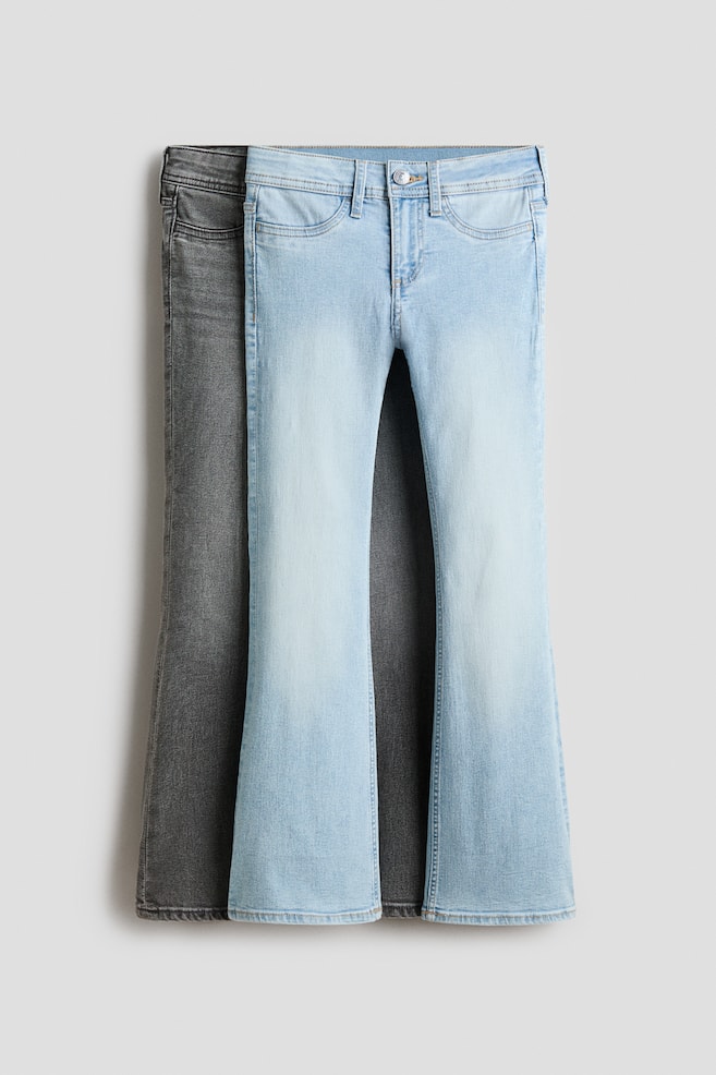 Two pairs of washed denim jeans are on display, with a front-facing light blue pair partially overlapping a dark grey pair. The light blue jeans feature a low waist, a zip fly with a button, and faux front pockets, while both pairs showcase a skinny fit through the hip and thigh that flares into wide legs. - 1