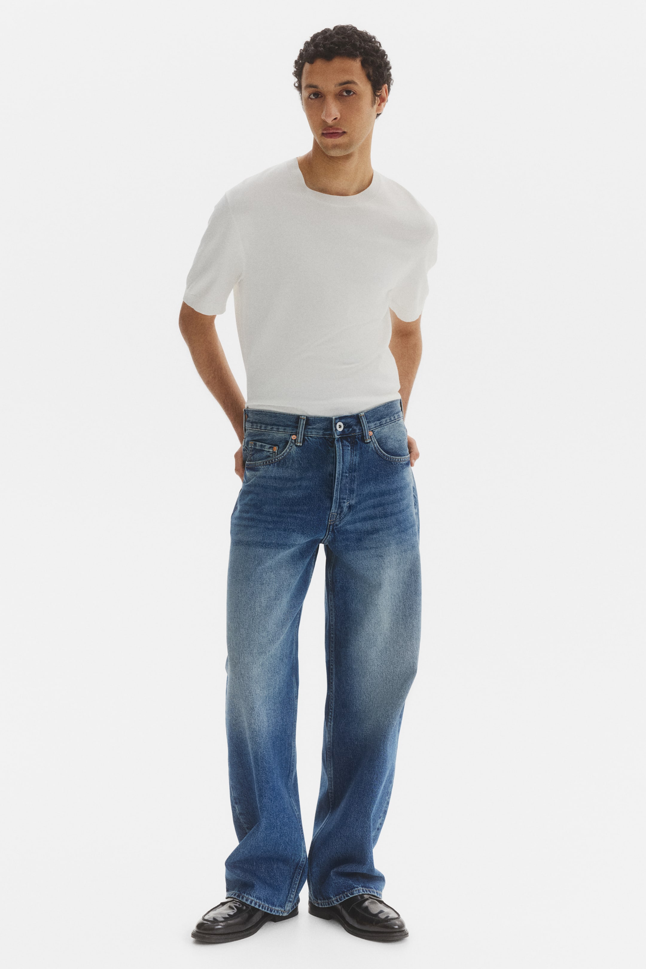 Größeres Bild ansehen: Blaue Jeans mit weitem Bein aus Denim präsentieren sich mit einem gewaschenen Finish und einem Knopfverschluss. Sie verfügen über Vordertaschen und einen lockeren Schnitt, mit einem weißen T-Shirt, das in den Bund gesteckt ist.