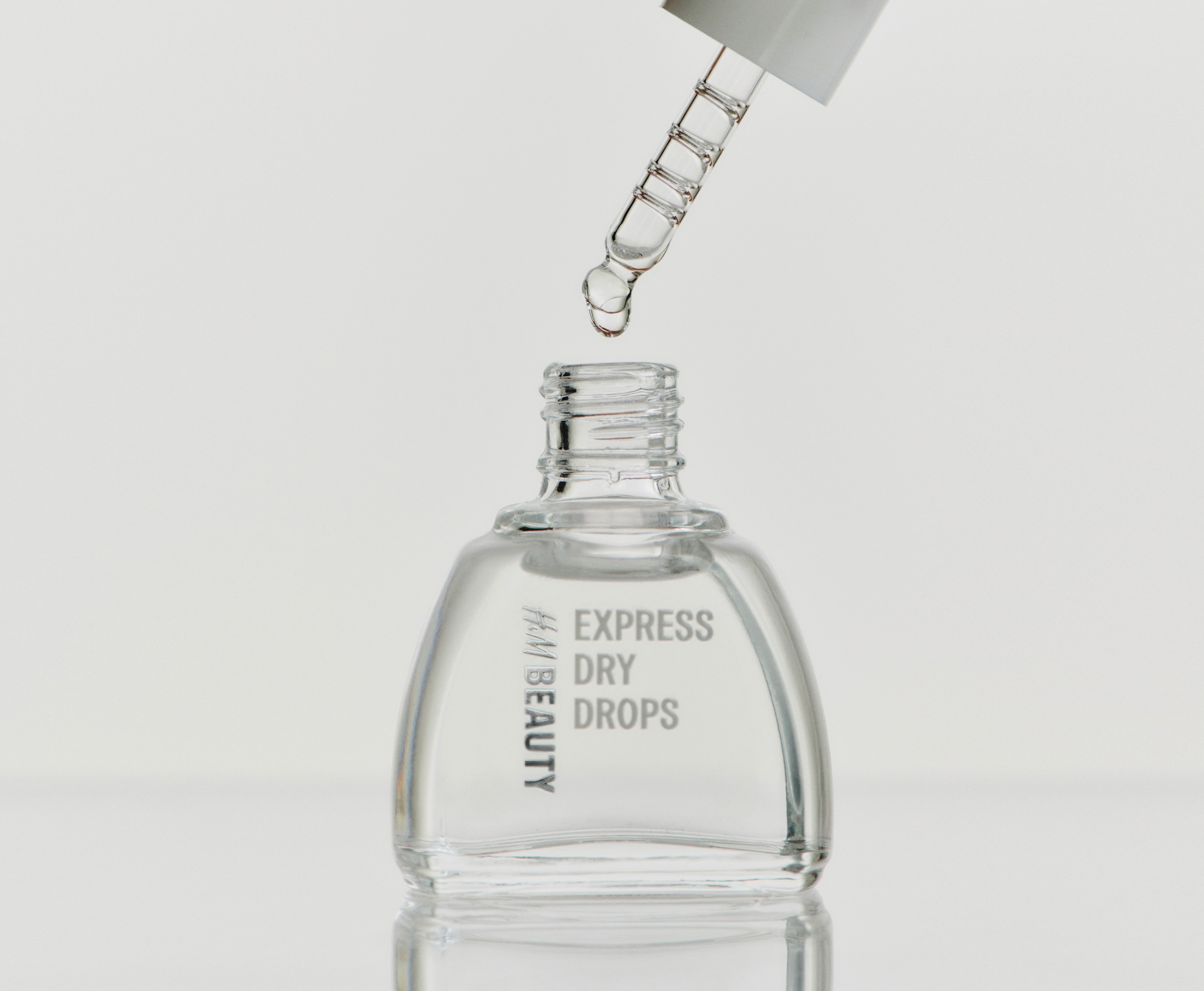 O sticlă cu picurător din sticlă transparentă, cu picături de uscare rapidă, este orientată frontal, prezentând un capac alb cu pipetă din sticlă vizibilă. Lichidul transparent umple sticla rotunjită, iar textul alb "H&M BEAUTY" apare vertical pe partea stângă, cu "EXPRESS DRY DROPS" apărând orizontal în centru.