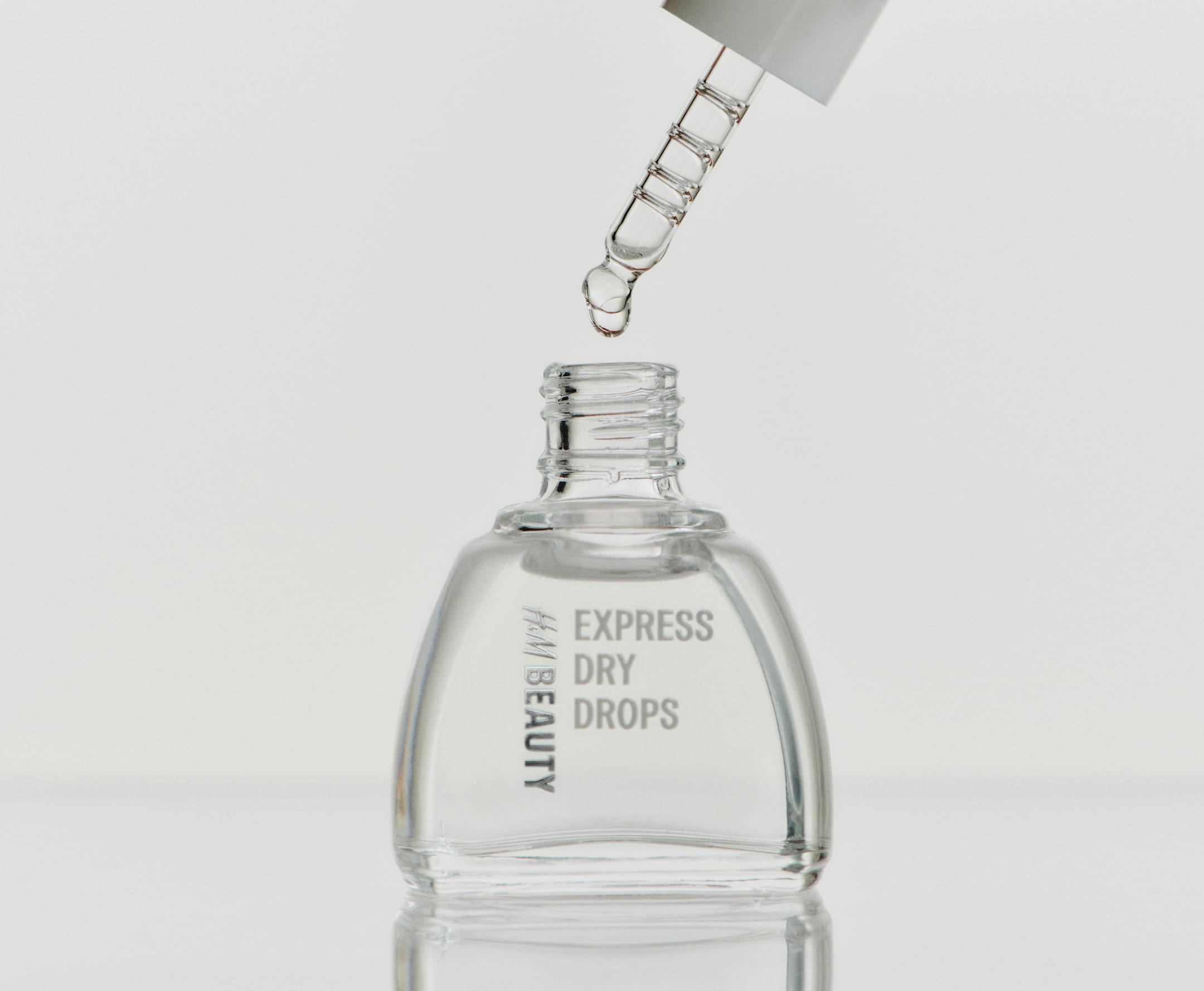 Größeres Bild anzeigen: Eine klare Glasflasche von H&M Beauty Express Dry Drops steht aufrecht und zeigt graue Markenaufschrift auf der Vorderseite. Ein Glastropfer schwebt direkt über dem geöffneten Flaschenhals und gibt einen einzelnen klaren Tropfen des flüssigen Produkts frei.