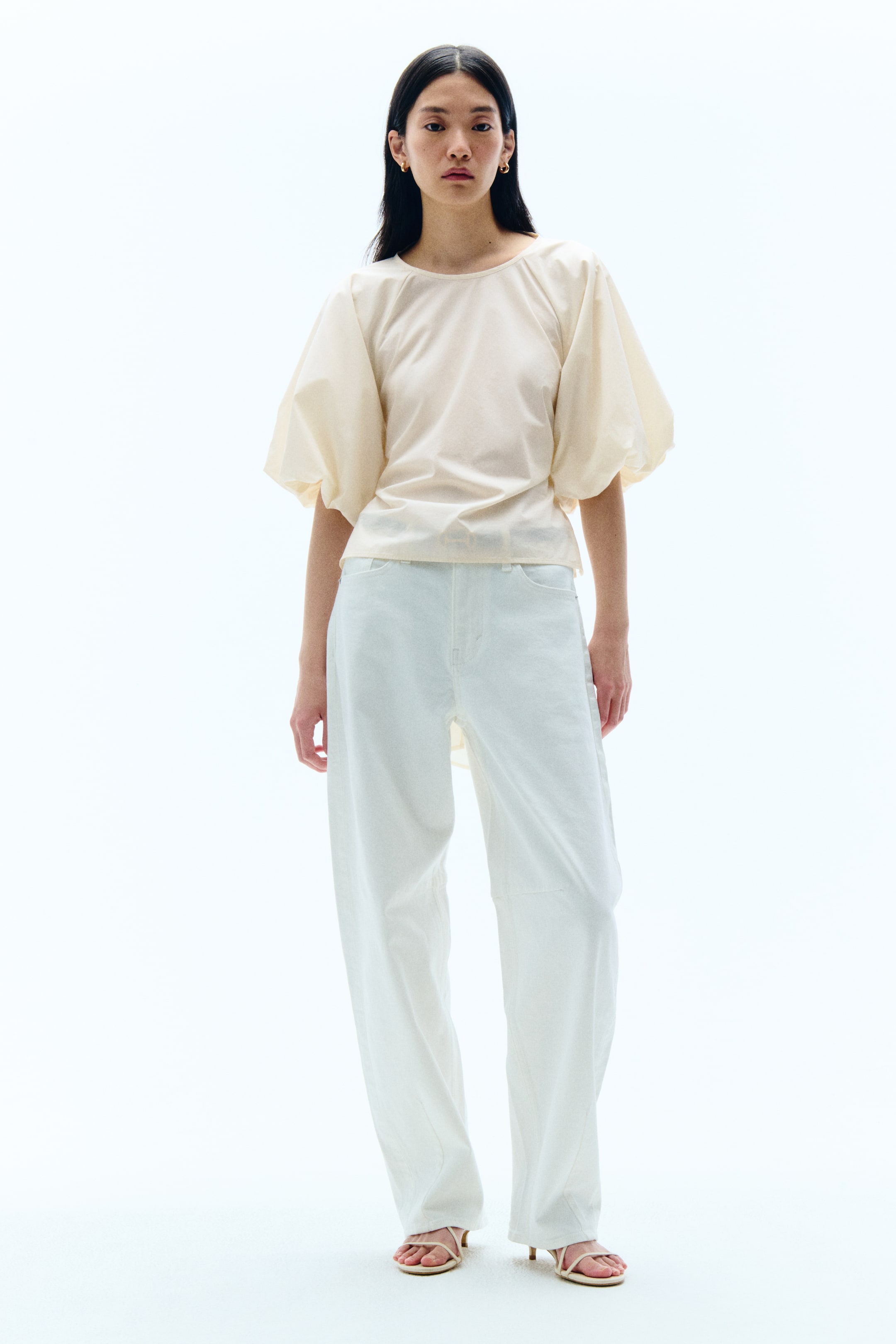 View larger image: A cream-coloured top, shown from the front, features a round neckline and voluminous elbow-length puff sleeves, gathered at the shoulders and cuffs. The top has a structured yet relaxed fit, with the front hem resting just above the waistline.