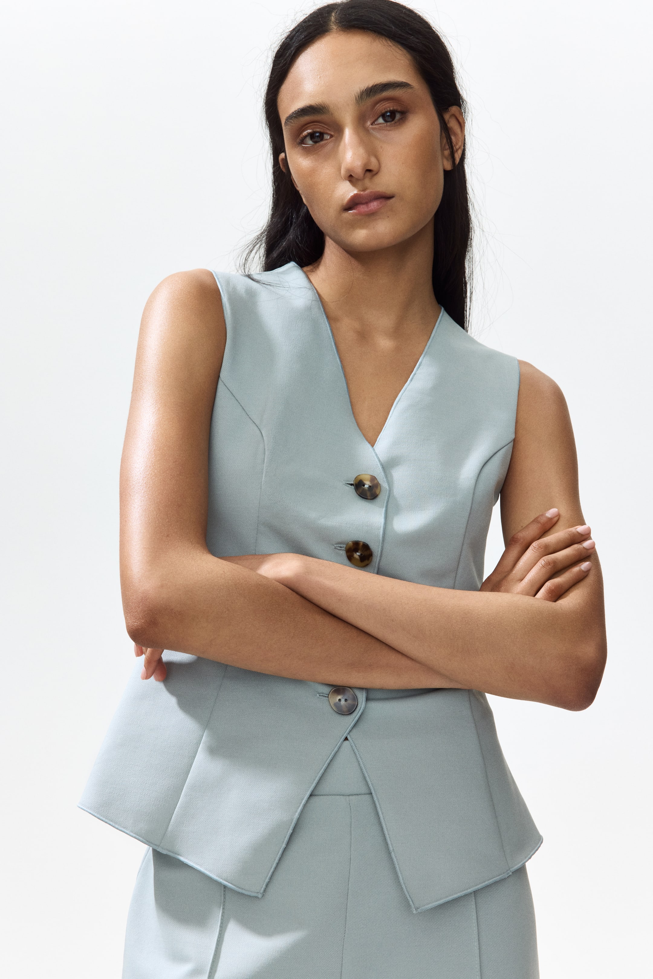 View larger image: A light blue sleeveless vest is donned, showcasing a V-neck and a button-front closure adorned with three brown tortoiseshell buttons. The vest boasts a flared hem with pointed front flaps, elegantly draping over coordinating light blue trousers, with a solitary grey button discernible near the lower flap.
