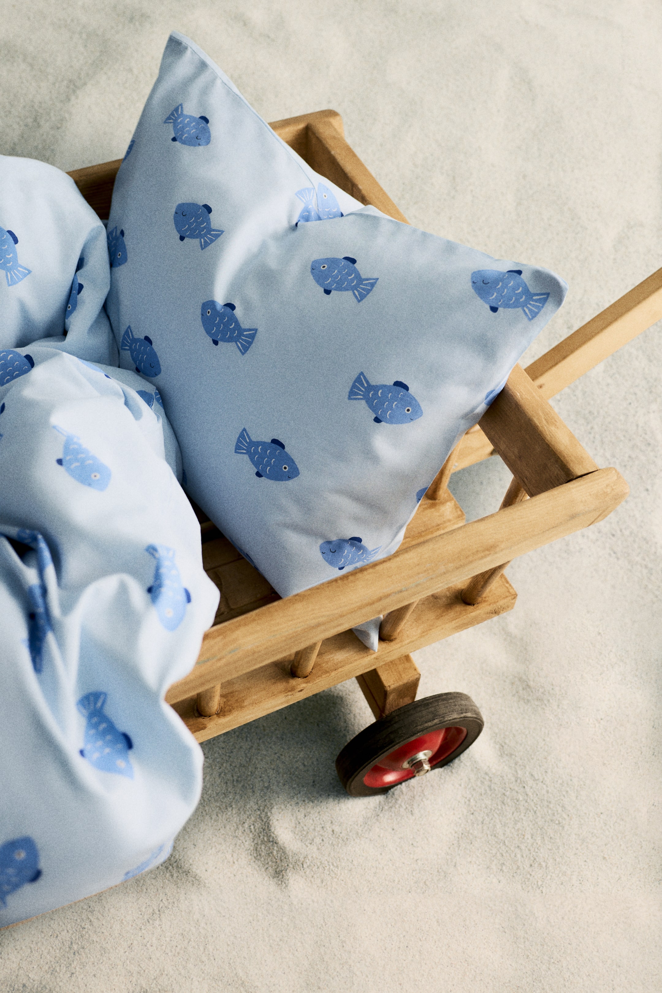 Vis større bilde: Lyseblått sengetøy, bestående av en pute og et sammenbrettet tekstil, viser et gjentakende mønster av mørkere blå fisker. Puten er vinklet litt oppover, og tekstilet er drapert ved siden av den i en liten trillevogn av tre.