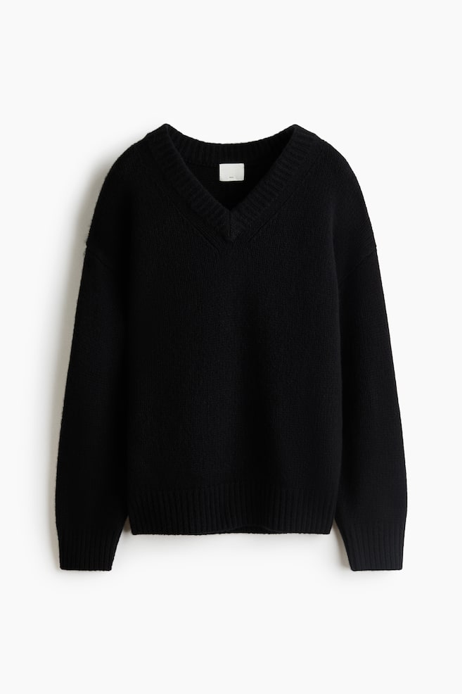 Un pull en laine tricotÃ©e noir et oversize est prÃ©sentÃ© de face, avec un col en V Ã  bord-cÃ´tes, des Ã©paules tombantes, ainsi que des manches longues avec poignets cÃ´telÃ©s et un ourlet cÃ´telÃ© assorti. - 1