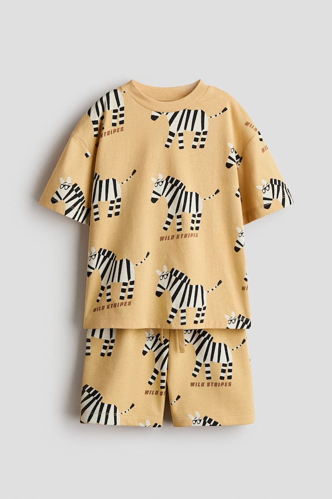 Ein hellgelbes Set mit Print, bestehend aus einem T-Shirt und passenden Shorts, zeigt seine Vorderansicht, verziert mit einem wiederkehrenden schwarz-weissen Zebra-Muster. Das T-Shirt präsentiert einen gerippten Halsausschnitt und überschnittene Schultern, während die Shorts mit einem elastischen Bund und Kordelzug ausgestattet sind. - 1