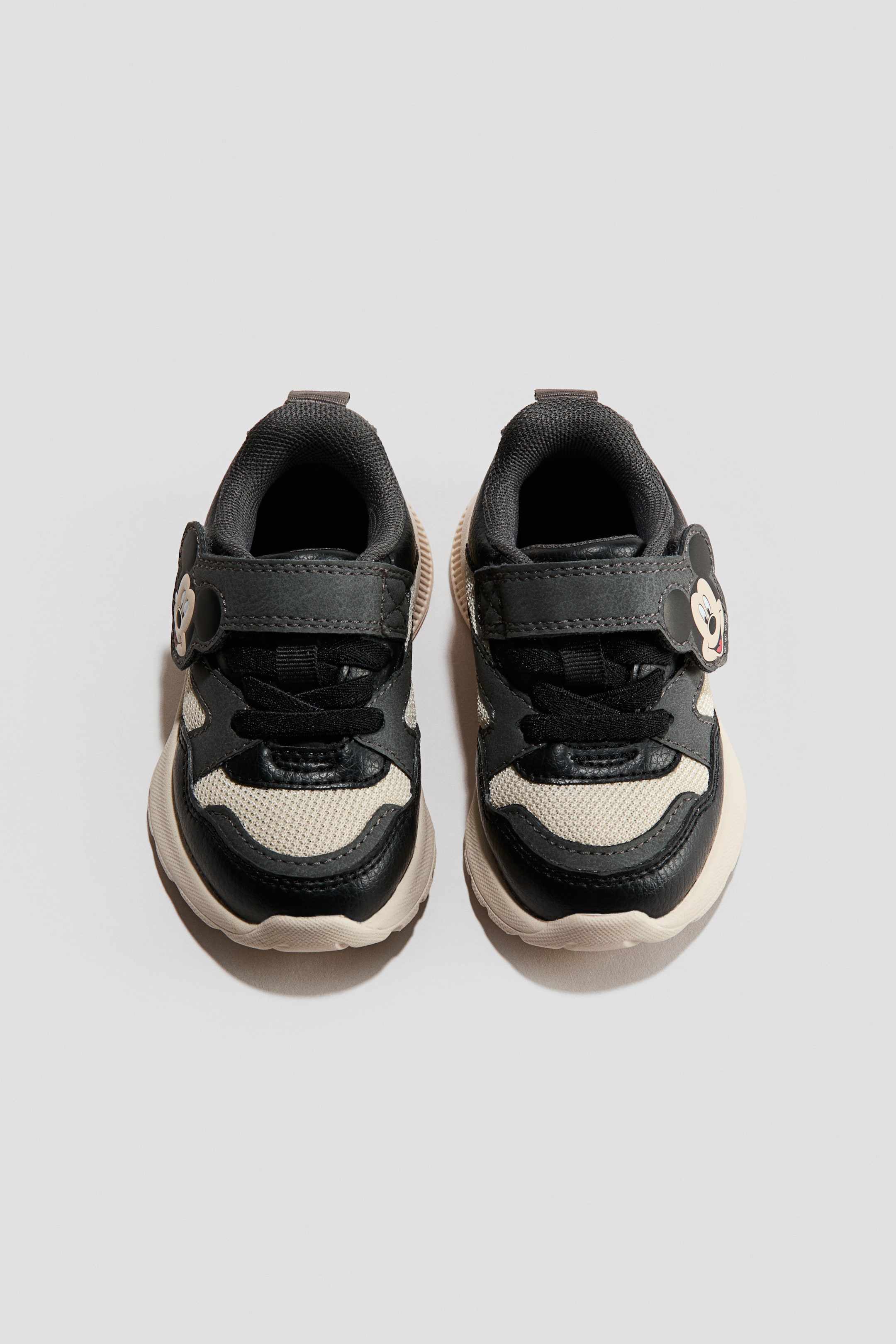 Visa större bild: Två barnsneakers är vända framåt och har paneler i mörkgrått, svart och off-white. Varje sko är försedd med svarta snören, en kardborrerem prydd med ett emblem föreställande Mickey Mouses huvud och en kraftig off-white sula.