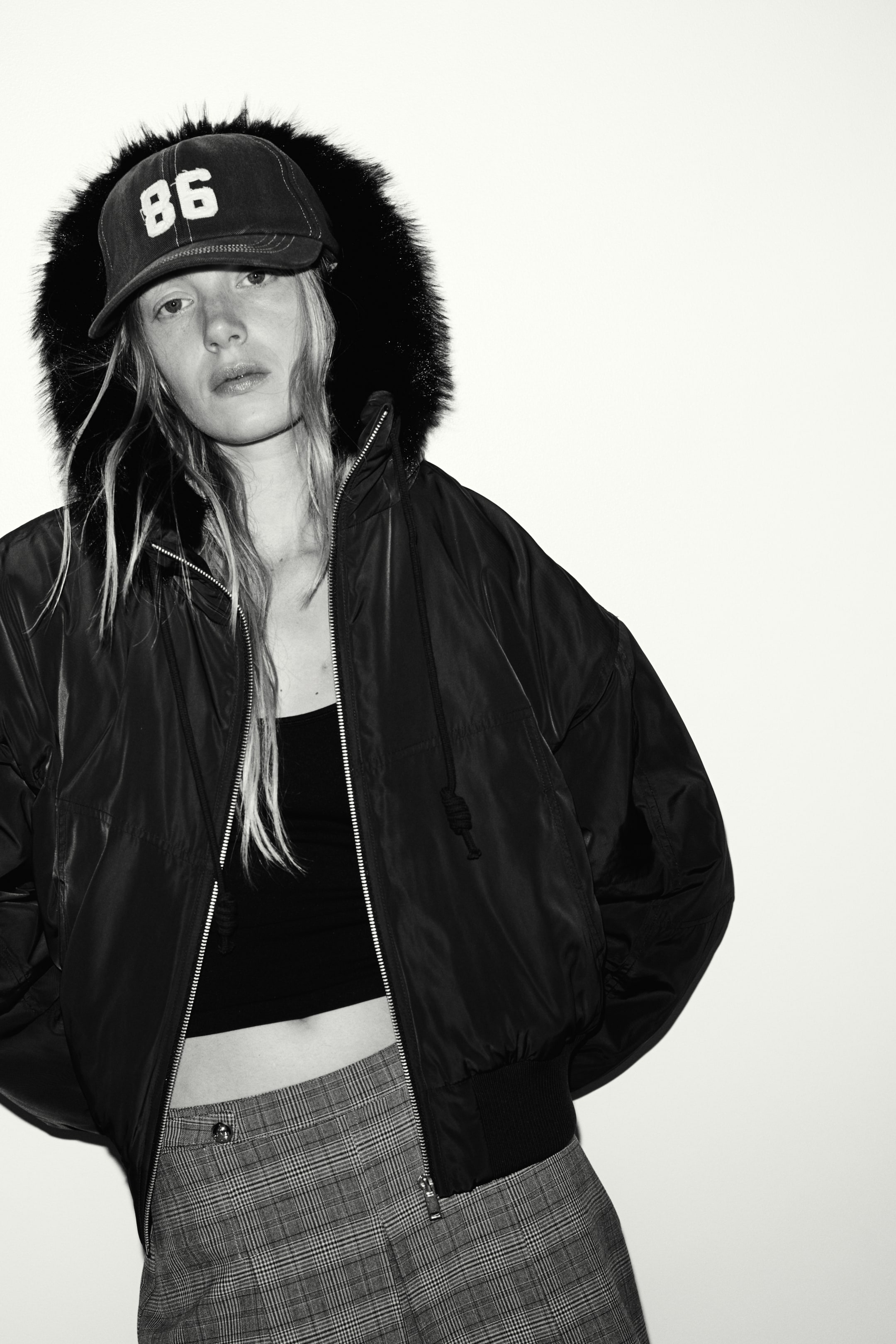 View larger image: An oversized, dark-coloured nylon jacket featuring a front zip, ribbed hem, and a drawstring hood with fluffy trim is being worn by the model. The jacket is styled open over a cropped top and plaid trousers, with the hood pulled up over a baseball cap.