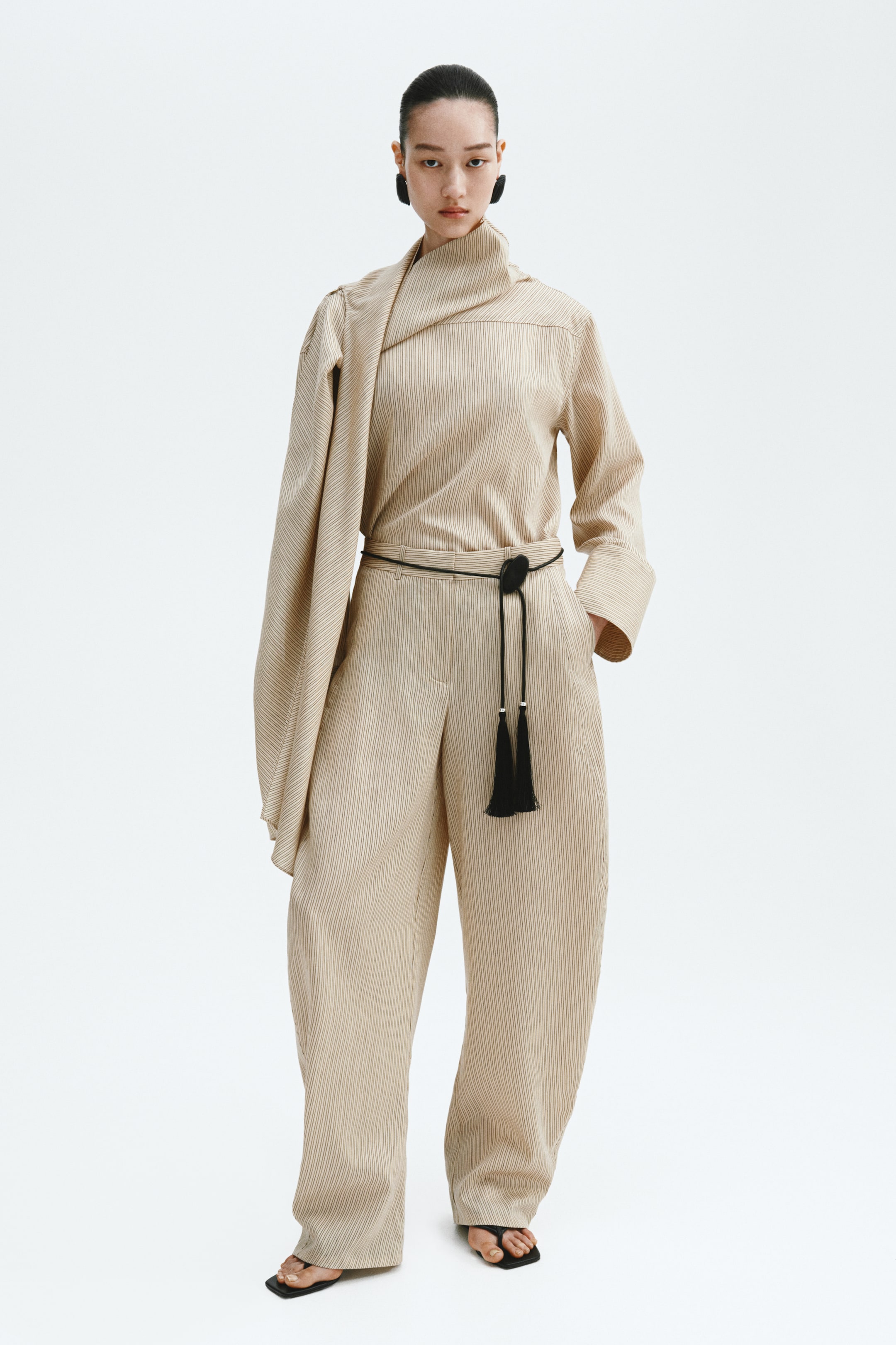 Ingrandisci l'immagine: Un completo a due pezzi a righe sottili di colore beige chiaro viene presentato, caratterizzato da un top a maniche lunghe con scollo drappeggiato alto e una manica drappeggiata sopra la spalla, insieme a pantaloni a gamba larga coordinati. Una sottile cintura a corda nera con un dettaglio ovale e due nappe stringe la vita.