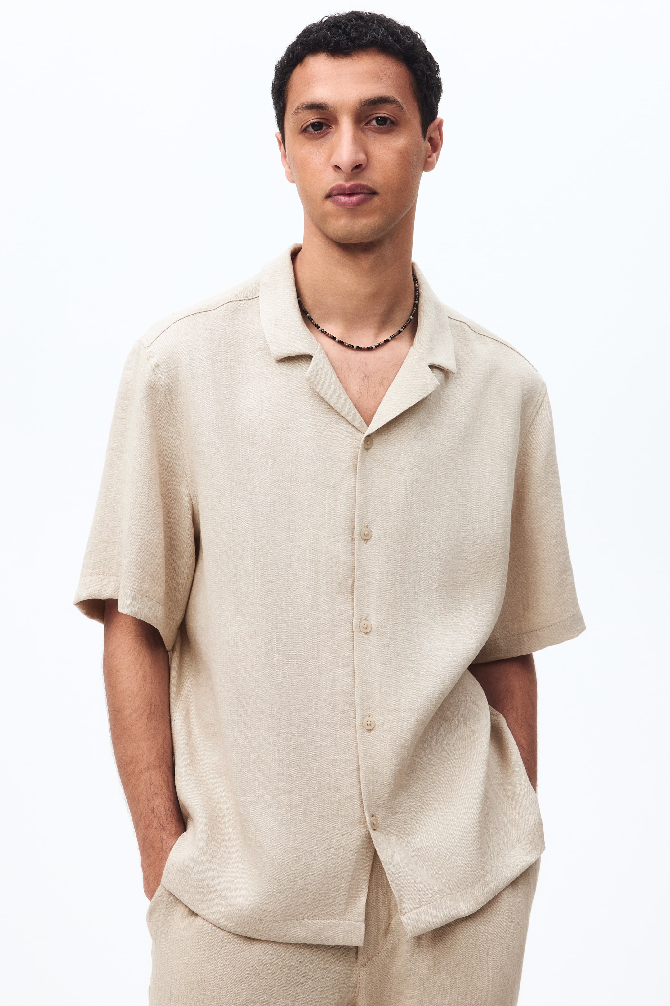 Xem ảnh lớn hơn: Áo sơ mi ngắn tay bằng vải dệt pha viscose chứa một ít linen, có cổ áo kiểu resort, hàng khuy giấu, gấu áo cắt thẳng và dáng rộng thoải mái cho một hình dáng giản dị, đang được người mẫu mặc. Người mẫu cũng đang đeo một chiếc vòng cổ đính hạt trên áo và quần đồng bộ.