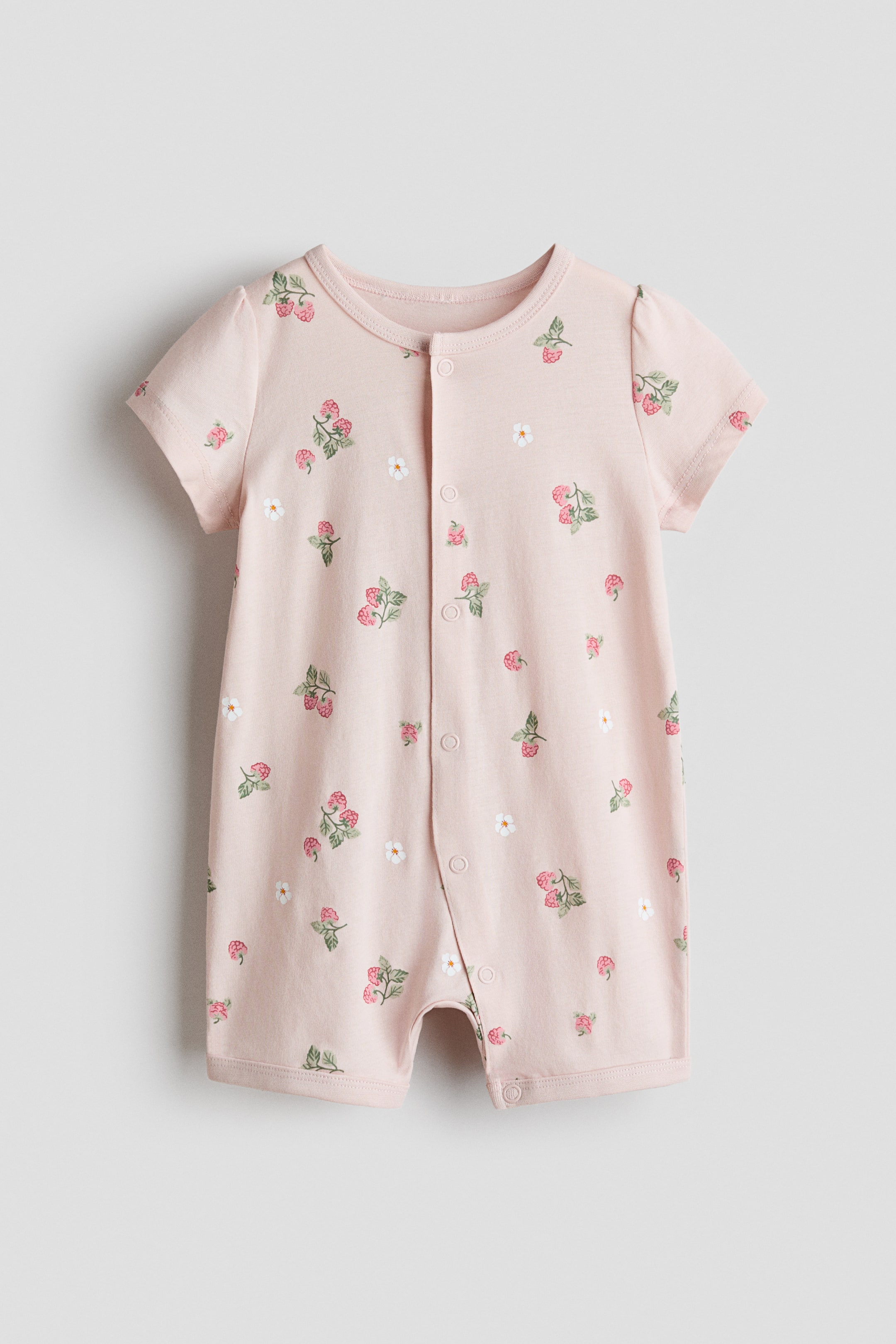 Agrandir l'image: Une grenouillère pour bébé est présentée de face, arborant une couleur rose clair avec un motif répétitif de framboises rouges accompagnées de feuilles vertes et de petites fleurs blanches. Elle est conçue avec des manches courtes, un col rond et des boutons-pression s'étendant sur le devant central.