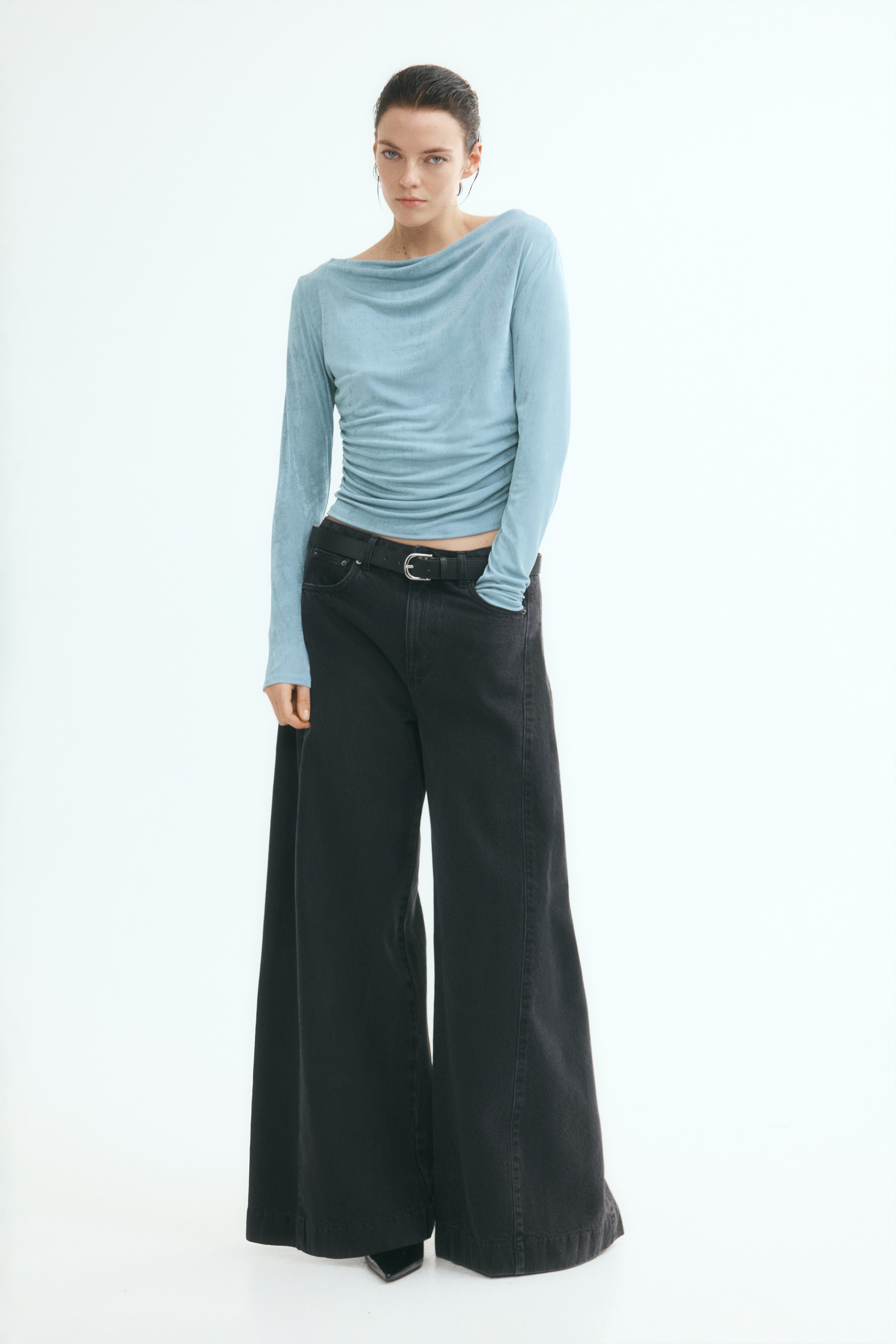 A light blue long-sleeve top is shown from the front, featuring a textured fabric and a draped boat neckline. The design includes ruching along the lower side seams, creating soft horizontal folds across the front hem.