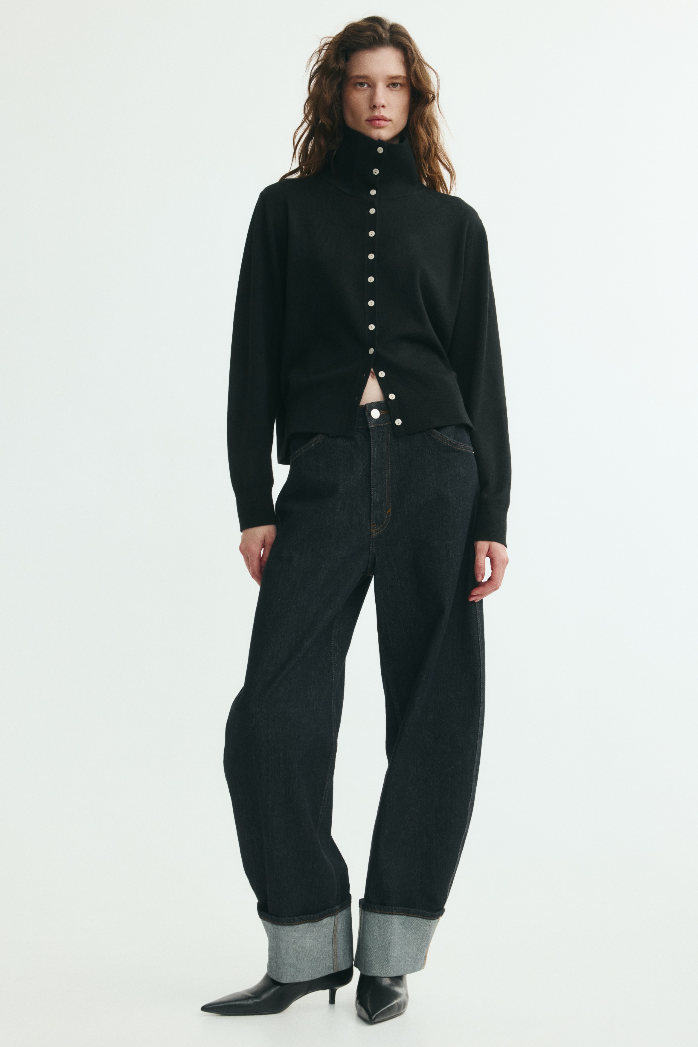 Een zwart, fijngebreid vest is van voren weergegeven en heeft een hoge hals, een volledige knoopsluiting aan de voorkant met contrasterende lichtgekleurde knopen, en lange mouwen. Het kledingstuk toont ribbeldetails aan de manchetten en zoom.