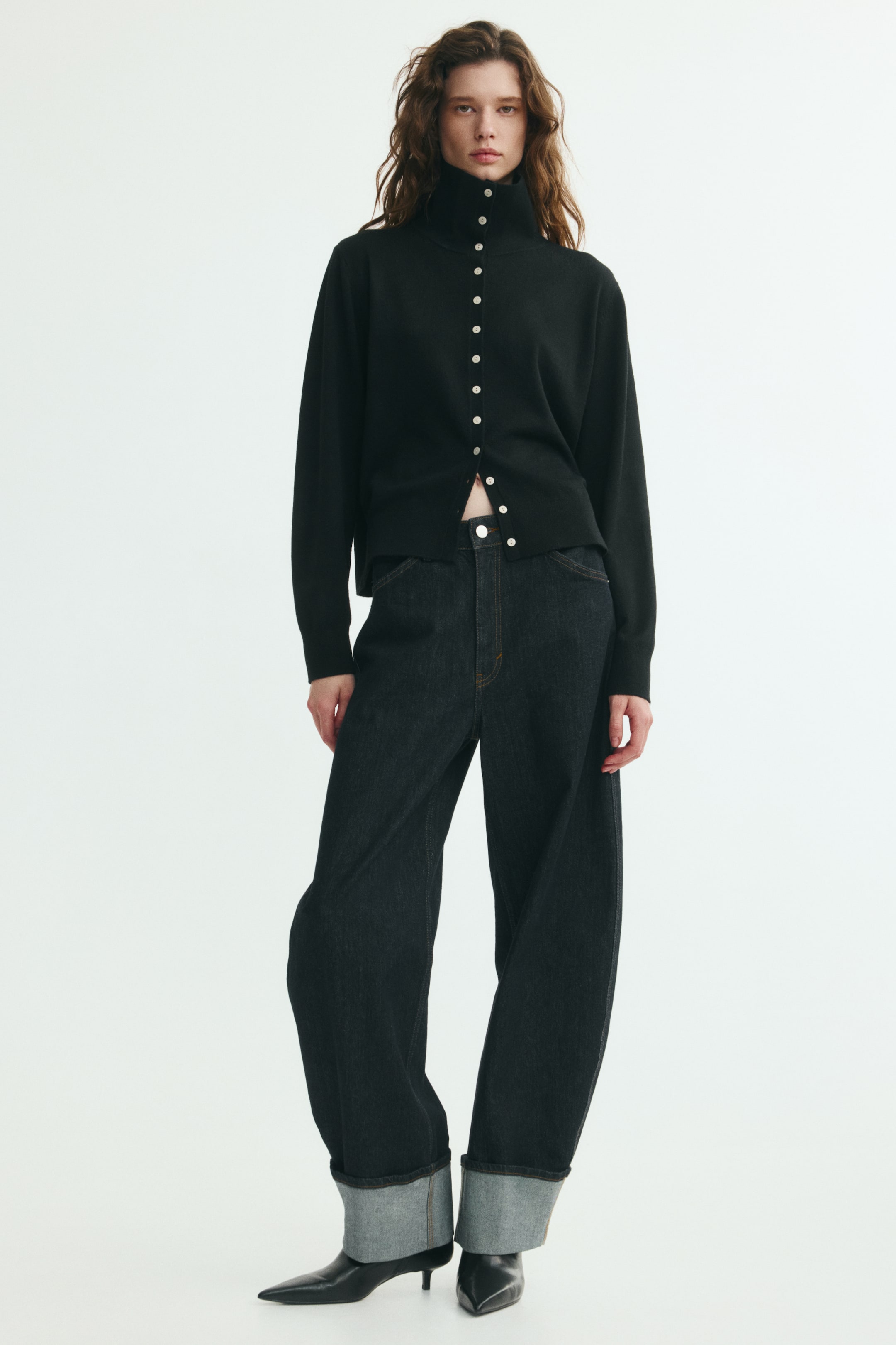 Grotere afbeelding bekijken: Een zwarte, fijngebreide cardigan met hoge hals, geribde boorden en contrasterende knopen aan de voorkant wordt gedragen door het model. De cardigan is gestyled met de onderste knopen open gelaten en gecombineerd met een wide-leg jeans in donkere wassing met omgeslagen pijpen.