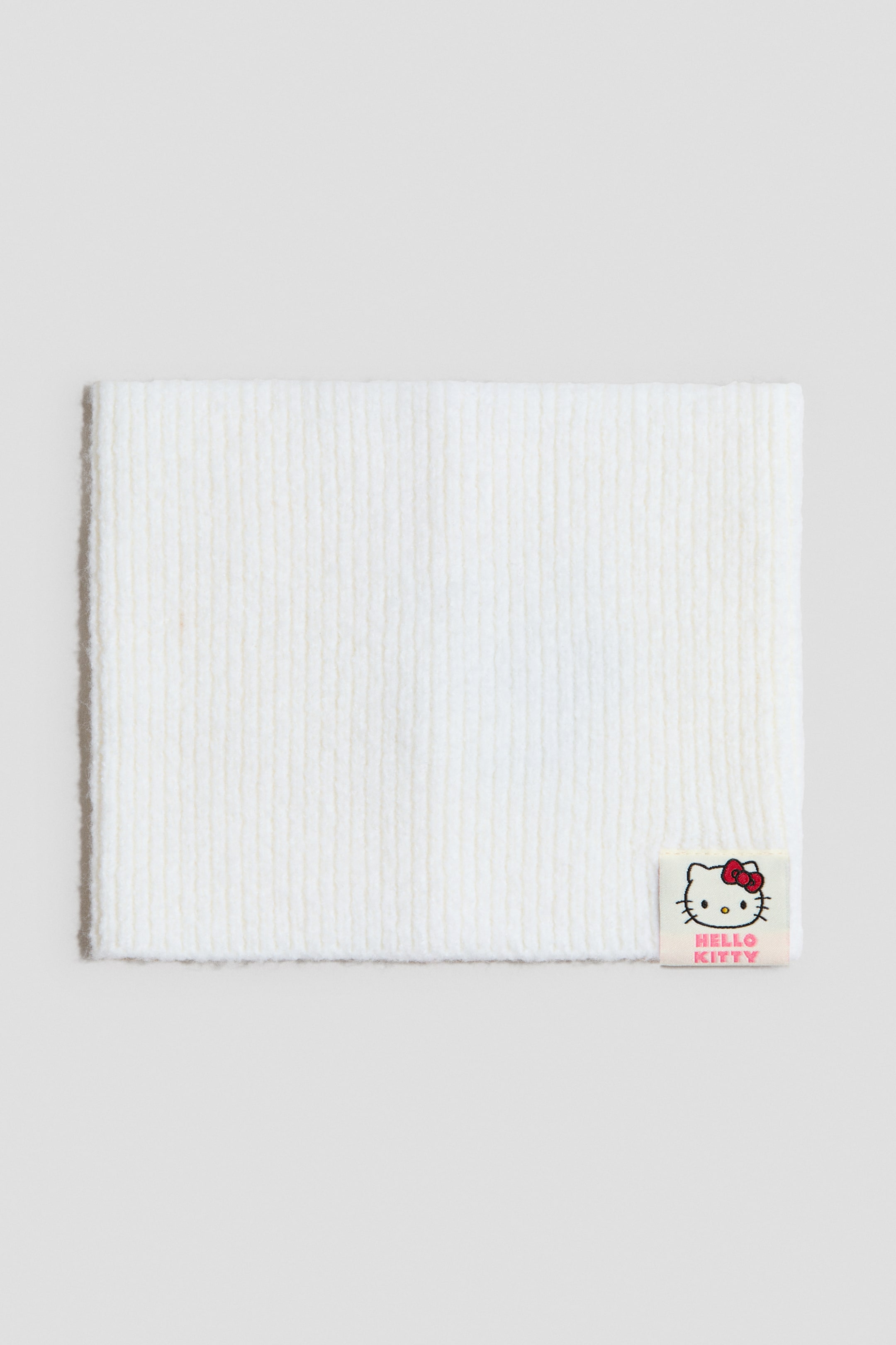 View larger image: A white knitted neck warmer is presented flat, showcasing a ribbed texture. A small square Hello Kitty logo tag is displayed on the front, positioned at the bottom right corner.