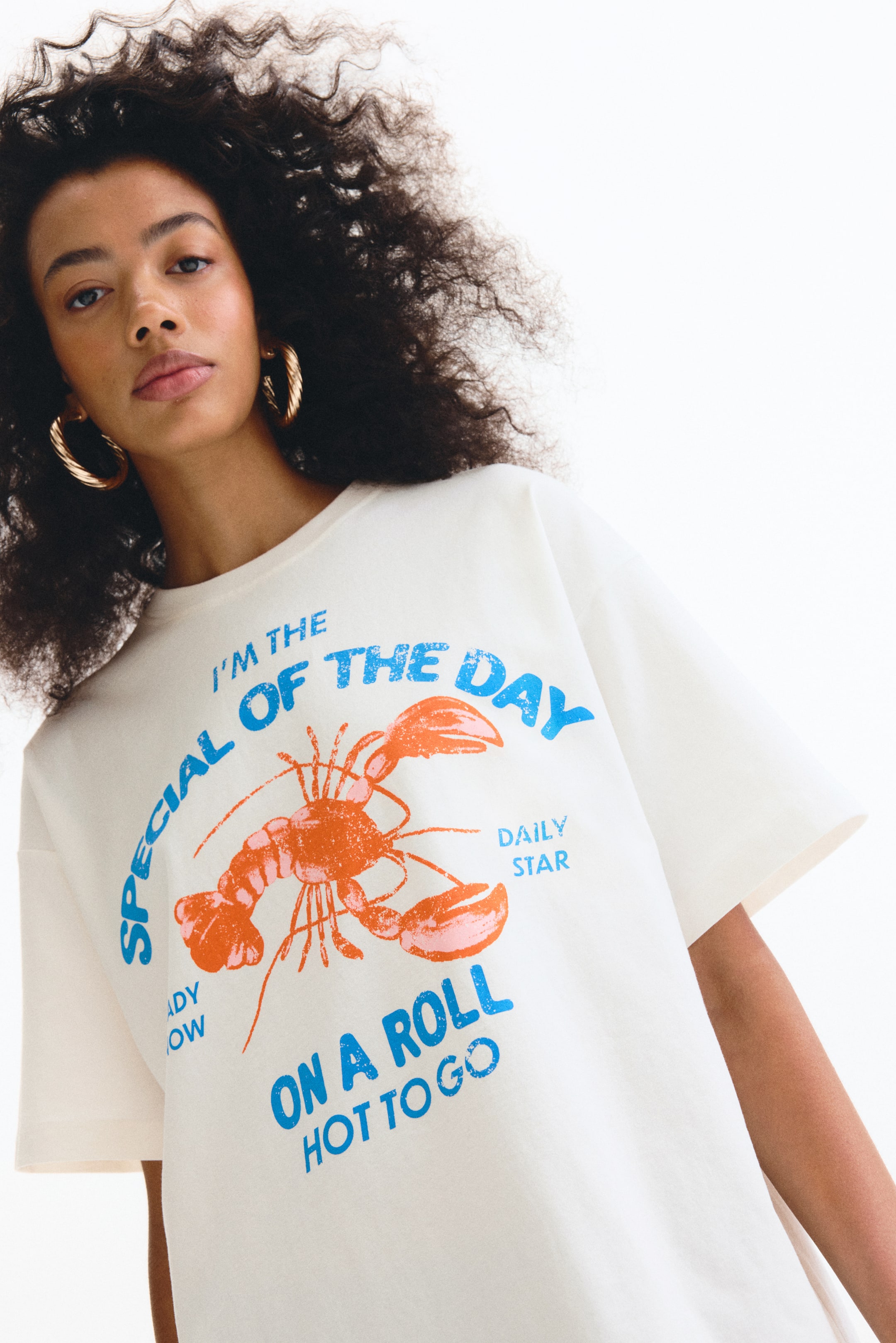 Größeres Bild ansehen: Ein off-white T-Shirt mit einem großen Grafikdruck auf der Vorderseite, das eine orangefarbene Hummer-Illustration zeigt. Über dem Hummer steht in blauer, abgenutzter Schrift "I'M THE SPECIAL OF THE DAY", während darunter "ON A ROLL HOT TO GO" zu lesen ist. Das T-Shirt hat eine entspannte Passform mit kurzen Ärmeln.