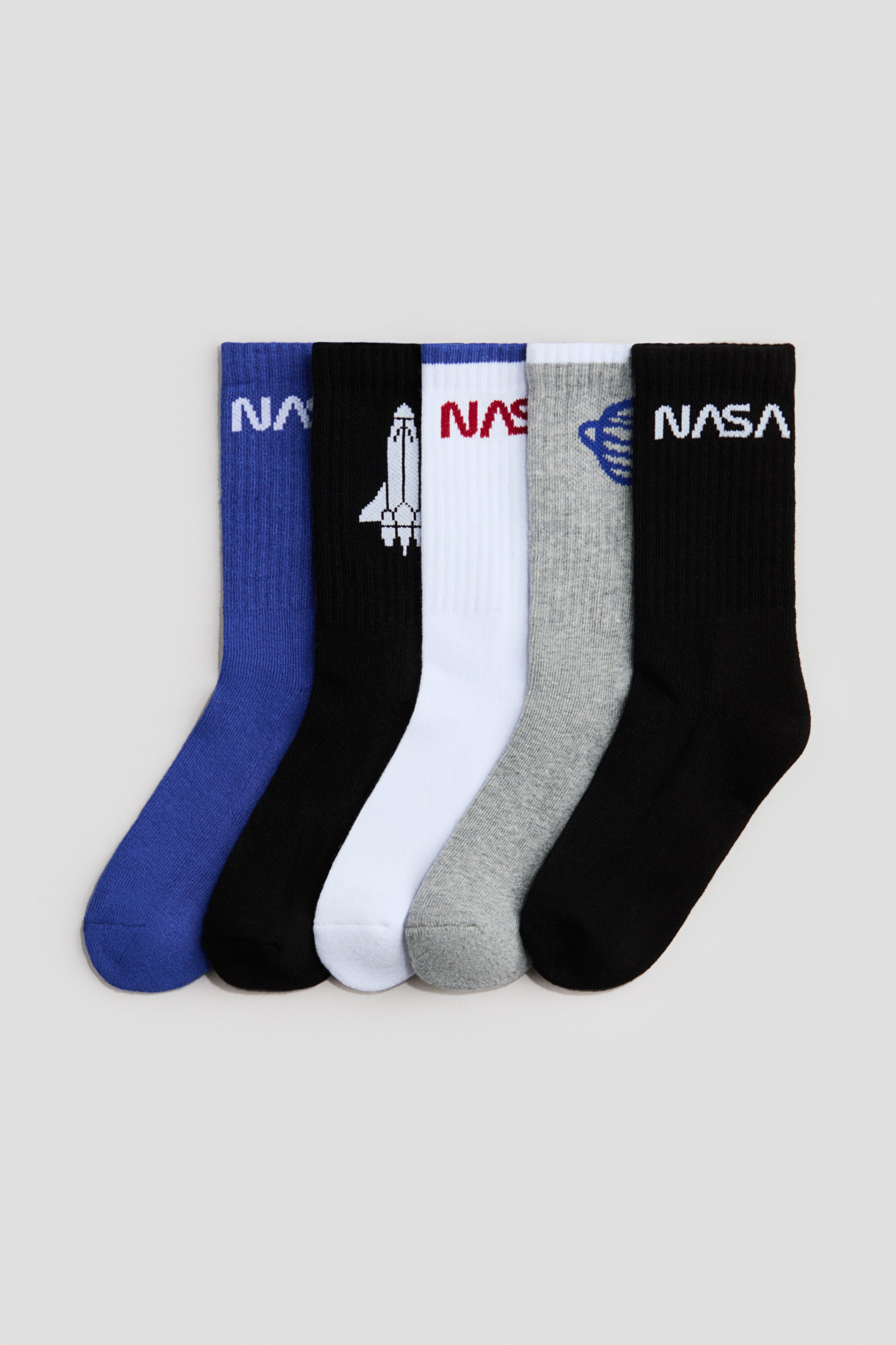 Ingrandisci l'immagine: Cinque paia di calze sono disposte in fila, presentando la parte anteriore. Presentano gambali a coste e vari design a tema NASA, inclusi testi e una grafica dello space shuttle. Le calze sono disponibili nei colori blu, nero, bianco e grigio.