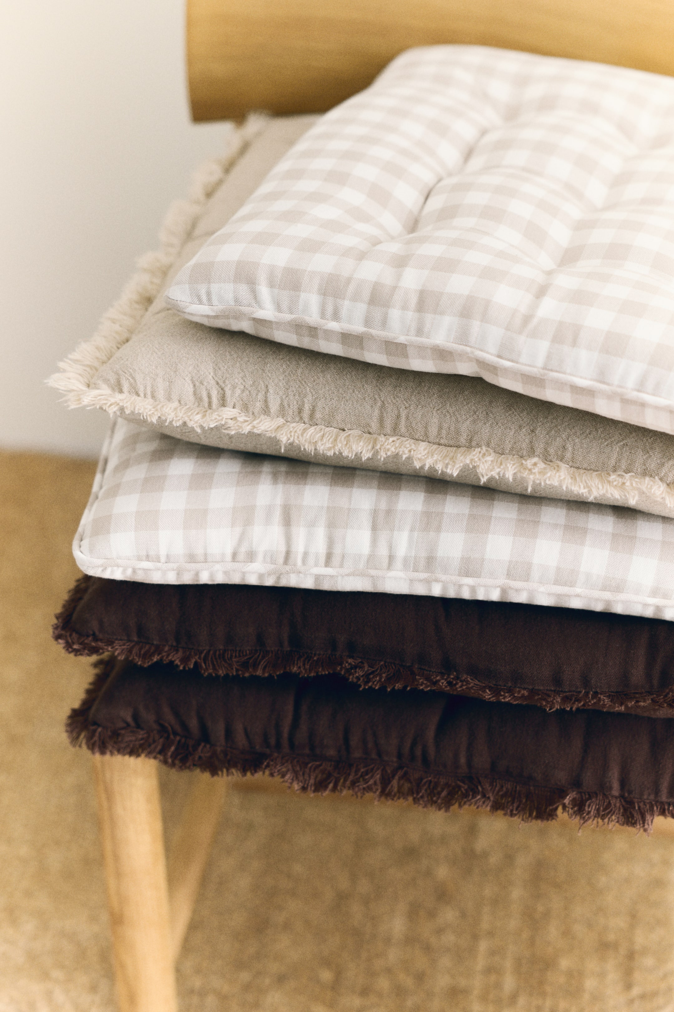 View larger image: Four seat cushions are stacked, each showcasing unique designs. The top and third cushions feature a light beige and white gingham pattern with tufted centers. The second cushion presents a solid light beige with frayed edges, while the bottom two cushions display a solid dark brown with frayed edges.