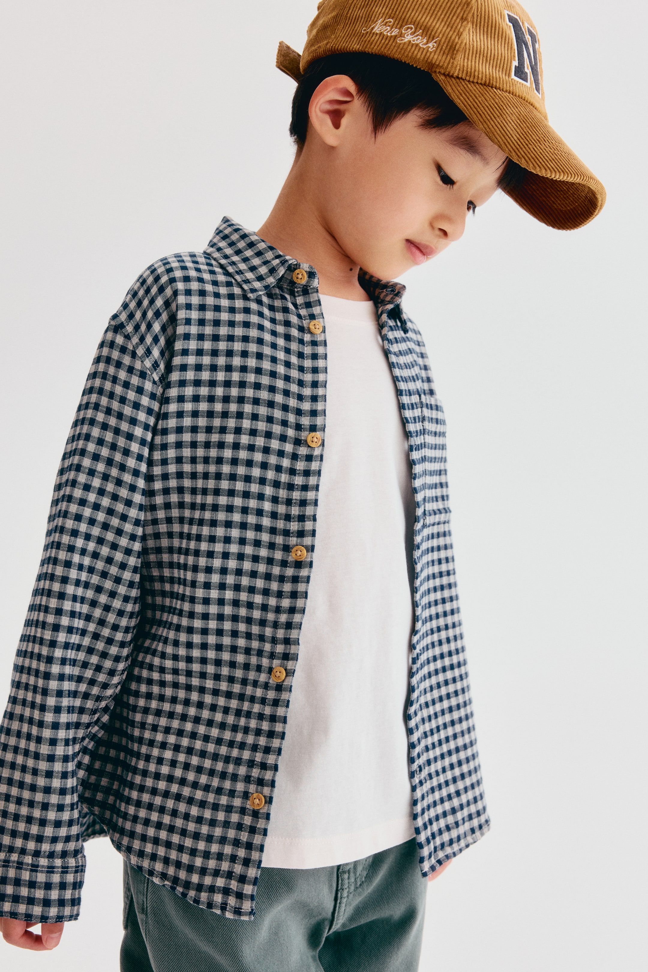 Grotere afbeelding bekijken: Een losvallend katoenen mousselineshirt in een marineblauw en wit gingham-ruitpatroon wordt open gedragen over een wit T-shirt, waardoor de ontspannen valling en de omgeslagen kraag mooi tot hun recht komen. Afgebeeld vanuit vooraanzicht, heeft het shirt lange mouwen met manchetten met knopen en contrasterende lichtbruine knopen langs de knoopsluiting aan de voorkant.