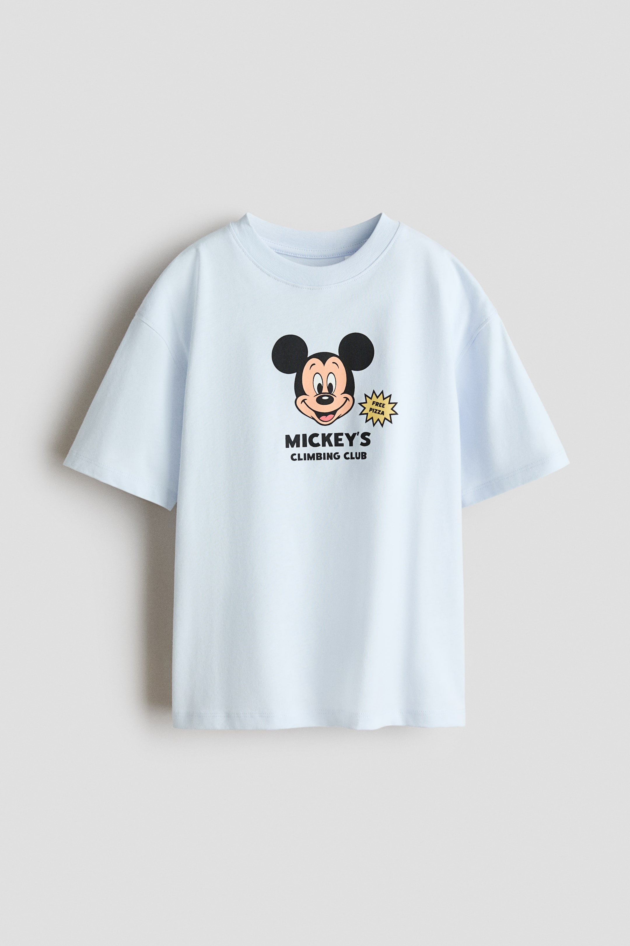 Visa större bild: En oversized t-shirt i mjuk, ljusblå bomullsjersey visas framifrån, med ribbad kant runt halsringningen och nedhasade axlar. Framsidan av t-shirten visar ett grafiskt tryck av Musse Piggs huvud, med texten "Mickeys Klätterklubb" under och en "Gratis Pizza" stjärnsprängning till höger.