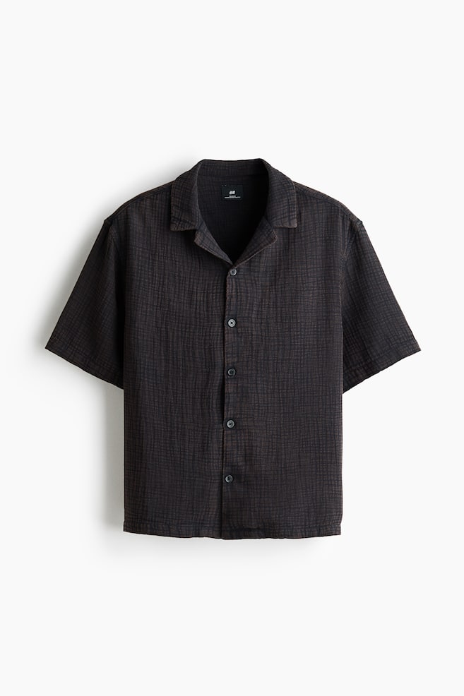A dark short-sleeved shirt is presented front-facing, featuring a textured, crinkled fabric with subtle grid lines. It is designed with a Cuban collar and a full button-front closure, secured with dark, round buttons. - 1