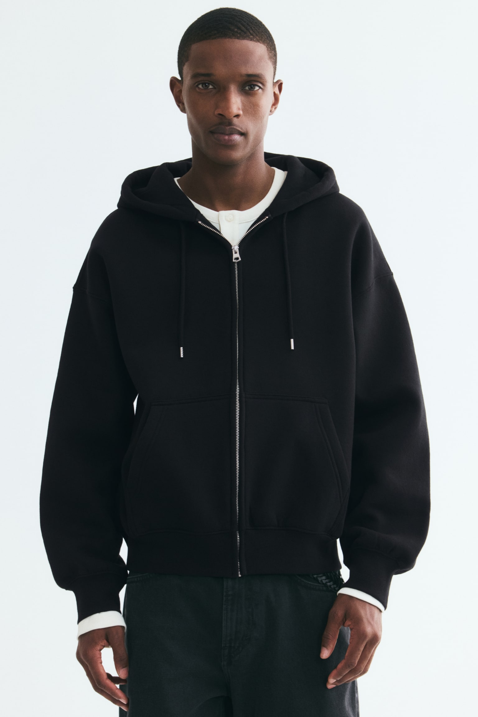 A black zip-through hoodie is showcased at the front, boasting an oversized fit with dropped shoulders. It's crafted with a full zip closure, a drawstring hood, long sleeves, front pockets, ribbed cuffs, and a ribbed hem. - 1