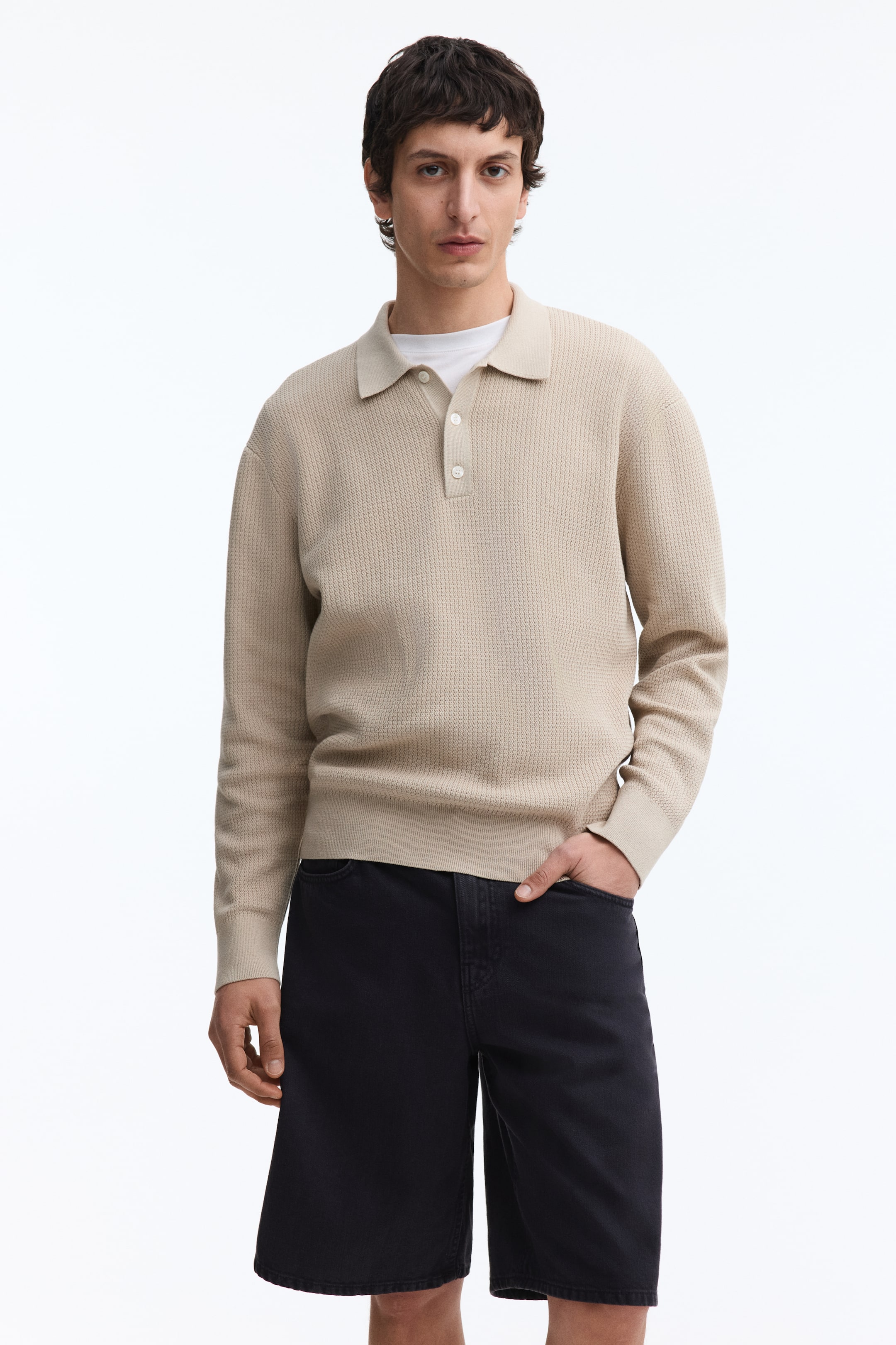 Grösseres Bild ansehen: Ein beige langärmliges Poloshirt wird von vorne gezeigt, mit einer strukturierten Strickoptik, einem Polokragen und einer Knopfleiste mit drei weißen Knöpfen. Es zeigt außerdem gerippte Bündchen und einen gerippten Saum, über einem weißen T-Shirt getragen.